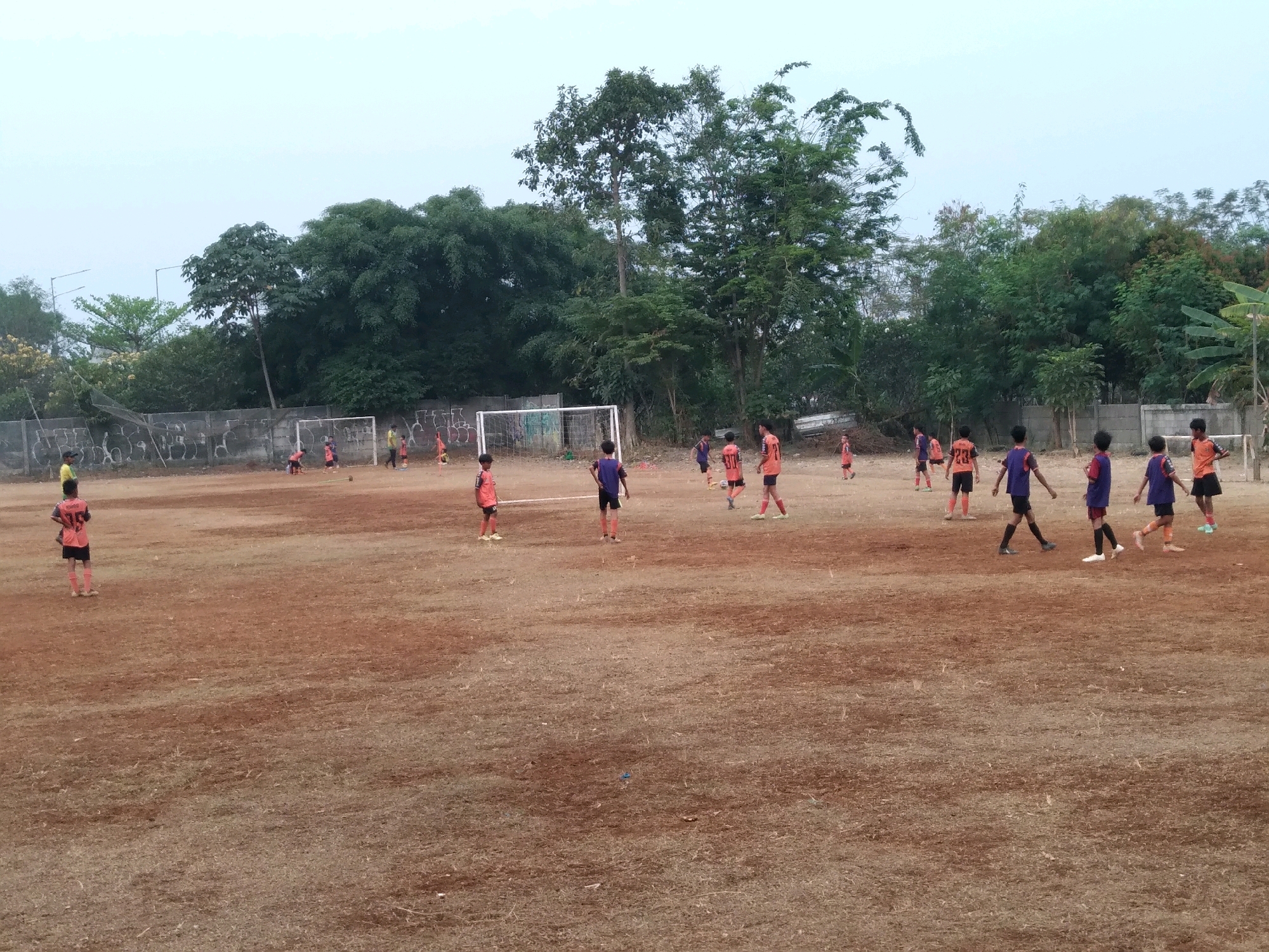 A group of young boys in soccer uniforms are on a dirt soccer field. Some are running, some are standing, and one appears to be kicking a ball. The field is dusty and uneven with patches of dry grass. In the background, there are trees, a fence with graffiti, and soccer goals. The sky is overcast and light. This scene appears to be in a semi-urban or rural area in South Tangerang, Indonesia, likely a local sports field or school ground.