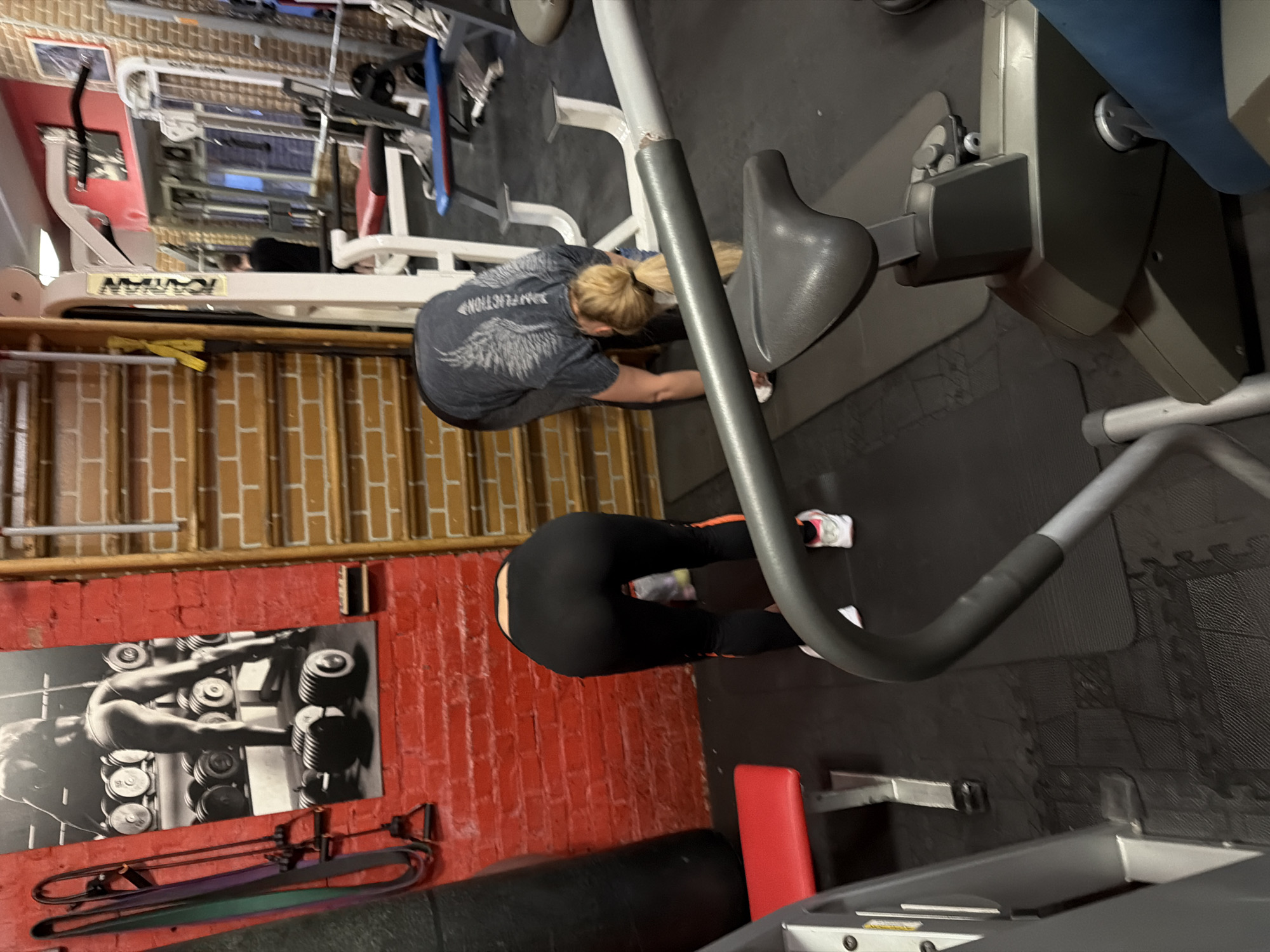 The image captures an indoor gym environment featuring two individuals engaged in stretching or exercise. The setting includes dark, interlocking rubber mats on the floor and a mix of red brick and exposed brown/orange brick walls.
In the mid-ground, a person with blonde hair tied in a ponytail is seen from behind, wearing a dark grey t-shirt with a white graphic of wings and possibly the word "PERFECTION" on the back, along with black leggings. This individual is bent forward from the waist, reaching towards the floor. To the right and slightly in the background, the lower half of another person is visible, also bent forward. This person wears black leggings with orange stripes along the sides and white sneakers with pink accents.
Various pieces of gym equipment are present. A stationary bike or similar cardio machine with a grey seat and handle is prominent on the right side. A red padded bench is visible in the lower right corner. Metallic structures and frames of other gym machines are scattered throughout. On the red brick wall, a large black and white poster depicts a person lifting dumbbells. Below this poster, several resistance bands are hanging on a rack. The brand name "KAYAN" is visible on a white piece of gym equipment in the background. The scene is illuminated by artificial indoor lighting.