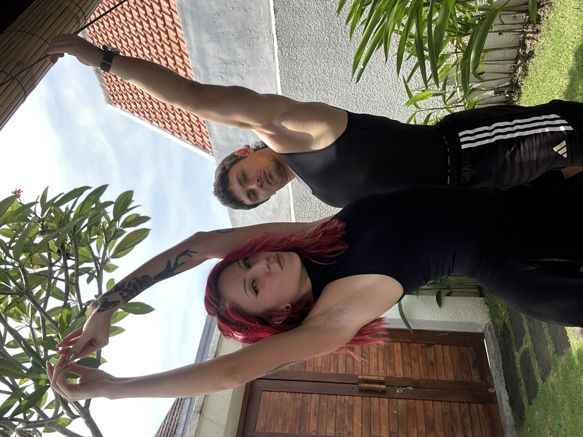 The image depicts a man and a woman in North Kuta, Indonesia, performing what appears to be a synchronized stretch. The woman, with red hair and tattoos on her arms, is closer to the camera and appears to be arching backward slightly with her arms raised overhead. The man, located behind her, also has one arm raised while the other may be pulling a cord. He is wearing a black tank top and Adidas shorts. The background includes a building with red tiled roof, trees, a wooden door, and grass. The time of day appears to be daytime, based on the lighting.