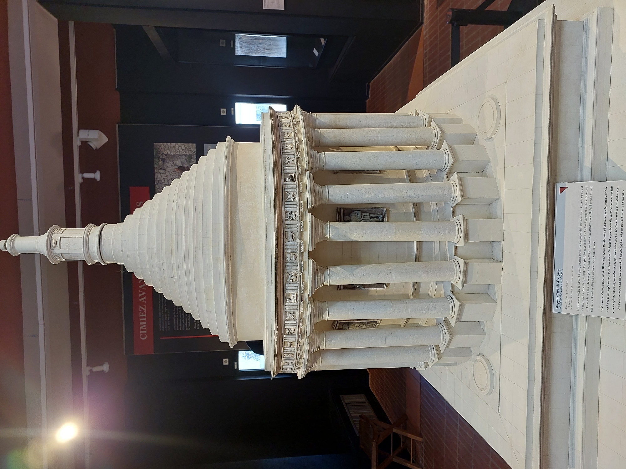 The image shows a detailed architectural model of the Trophy of Augustus, an ancient Roman monument located in the French Riviera city of Nice. The model is white and exhibits a circular structure with a tiered, conical roof topped by a finial. A colonnade of Corinthian-style columns encircles the base of the structure, and within the spaces between the columns, small relief sculptures are visible. The model rests on a multi-tiered white platform, and a descriptive plaque is affixed to the front of the base, providing historical information. The background suggests an indoor museum setting, with display cases, lighting, and other exhibits.