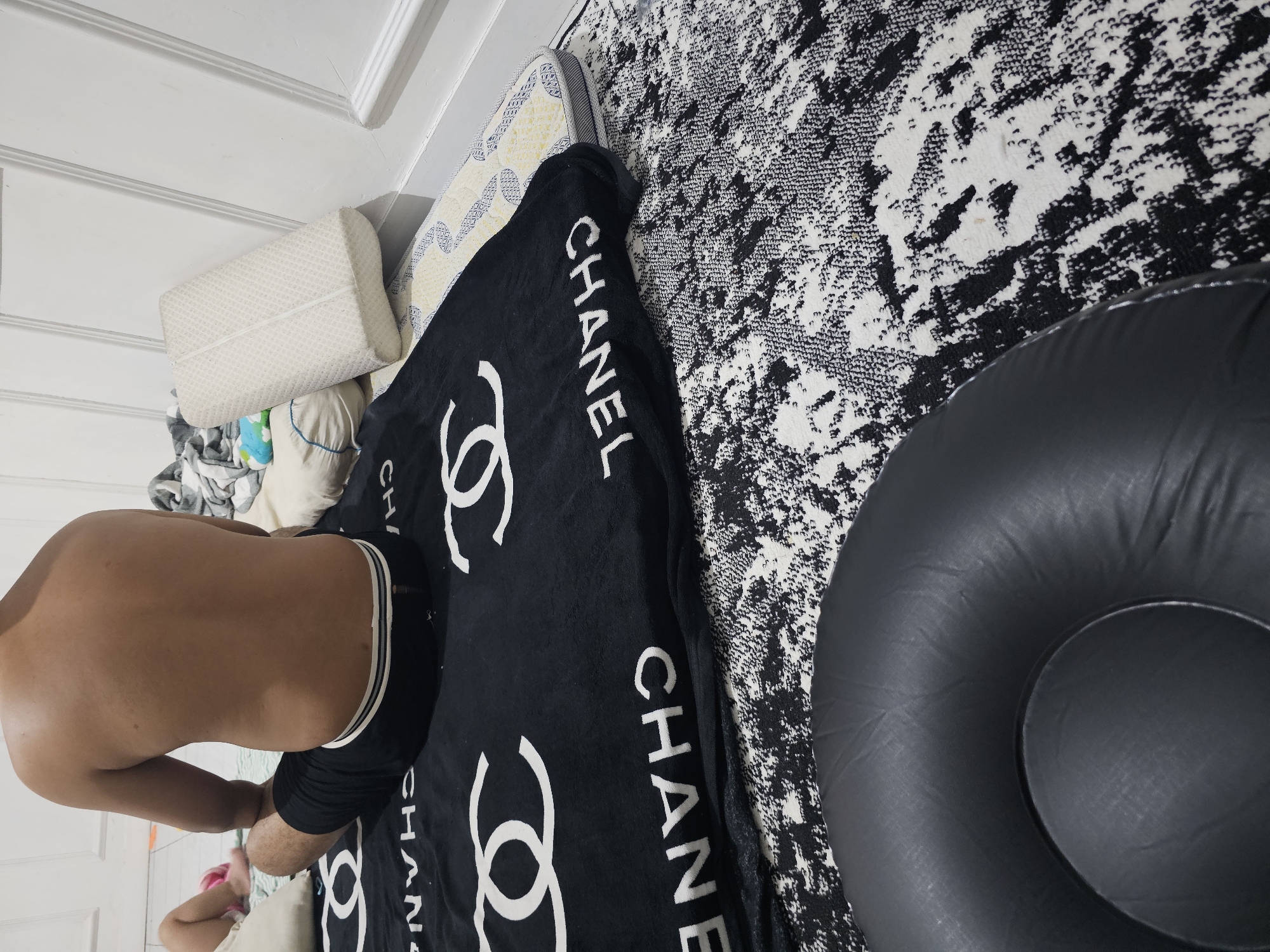 The image shows a person sitting on a Chanel blanket in what appears to be a bedroom in Lamongan, Indonesia. The person is bare-chested and wearing black shorts. The blanket is black with white Chanel logos and lettering. The person is sitting on a black and white patterned rug. In the foreground, there is a black inflatable ring. In the background, there is a white wall with molding, and a few pillows. The lighting is dim.