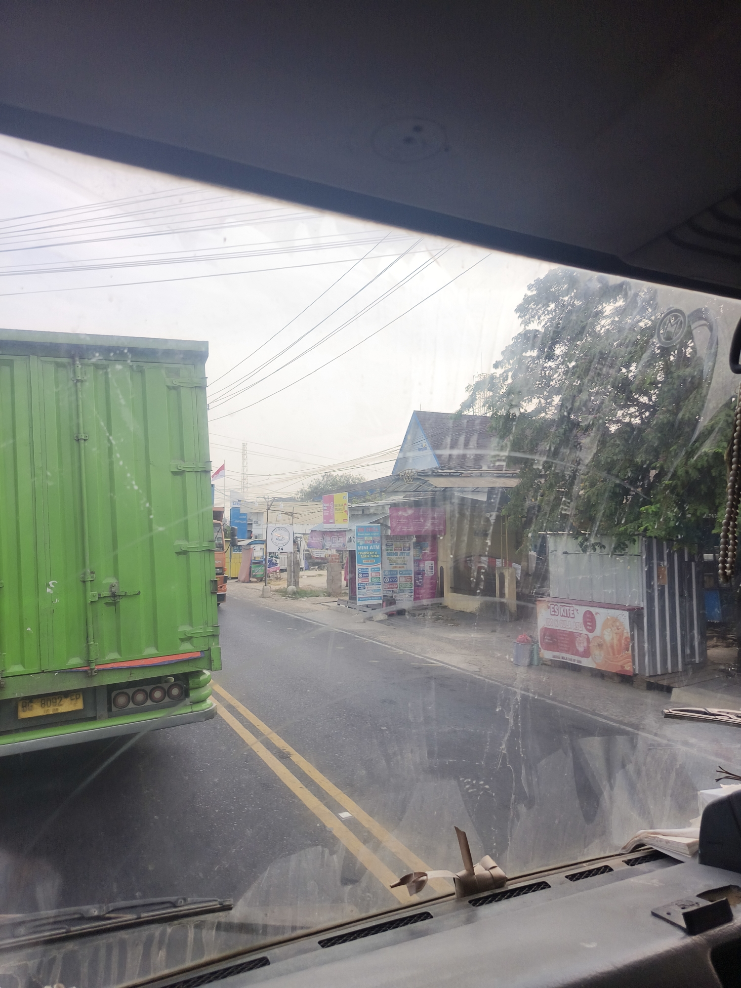 The image provides a view from inside a vehicle looking out through the windshield at a roadside scene in Pangkalpinang, Indonesia. On the left, a large green truck container is prominently visible, taking up a significant portion of the frame. The truck has a license plate that reads "BG 8092 EP". The road in front is asphalt with double yellow lines indicating traffic direction.
To the right of the road, there are several buildings and businesses. One building has a blue and gray roof. There are various signs and advertisements displayed, including one for "ES KITE" with images of drinks. A small, light-colored container is placed near this sign. Trees with dense foliage are visible behind the buildings, and power lines crisscross the sky. The overall atmosphere appears to be overcast. A few small objects, possibly debris or dirt, are visible on the windshield. The dashboard of the vehicle is partially visible at the bottom of the frame.