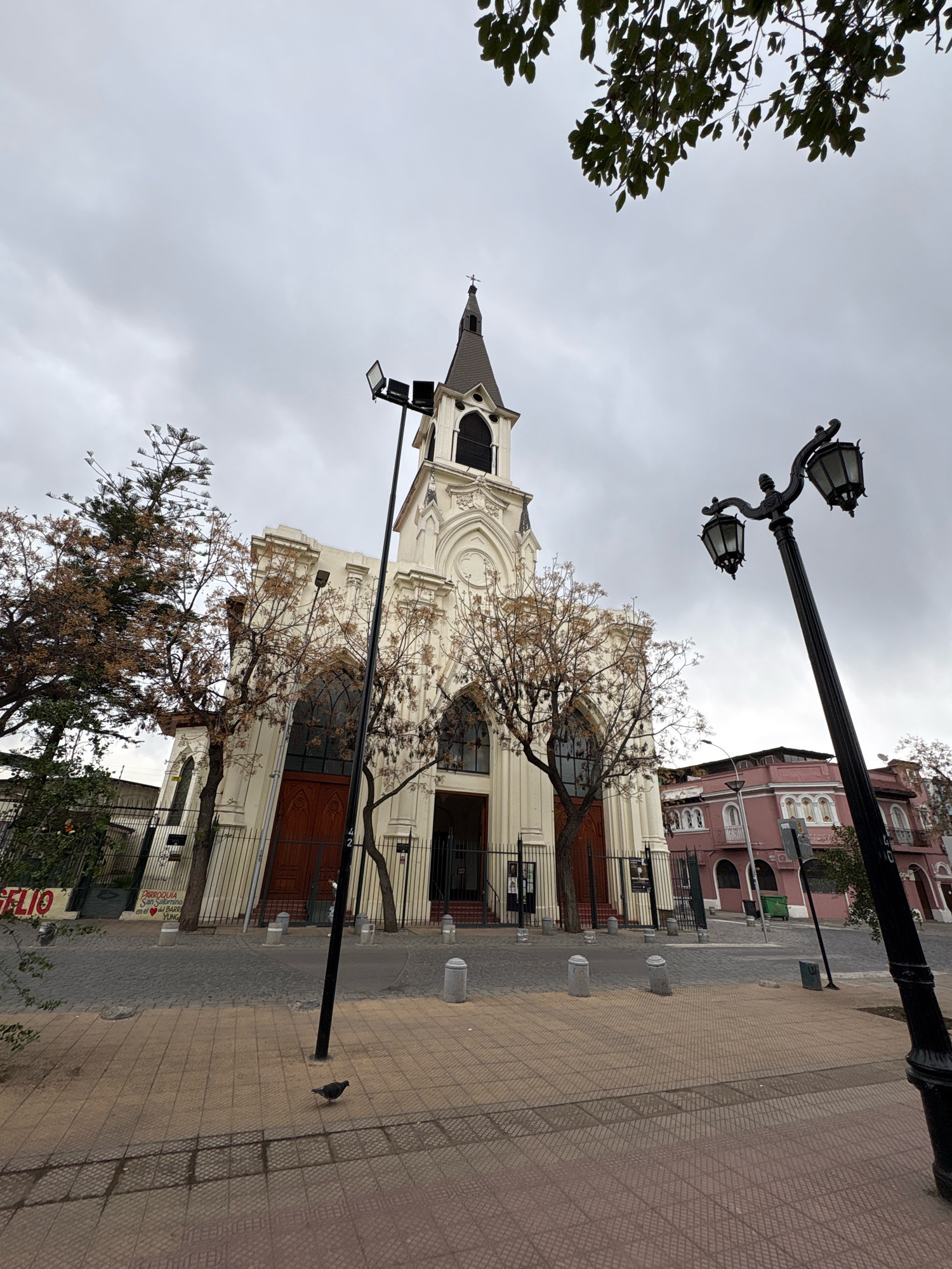 The image shows a view of the Church of San Vicente Ferrer in Santiago, Chile, on a cloudy day. The church, a white building with a tall spire and arched windows, is in the background. In front of the church is a paved sidewalk and street. A black lamppost with two lights stands in the foreground. A pigeon is walking on the sidewalk. Trees with bare branches are on either side of the church. A pink building is visible to the right of the church. The street in front of the church is paved with cobblestones. A sign on the left side of the church reads "SAN FELIO."