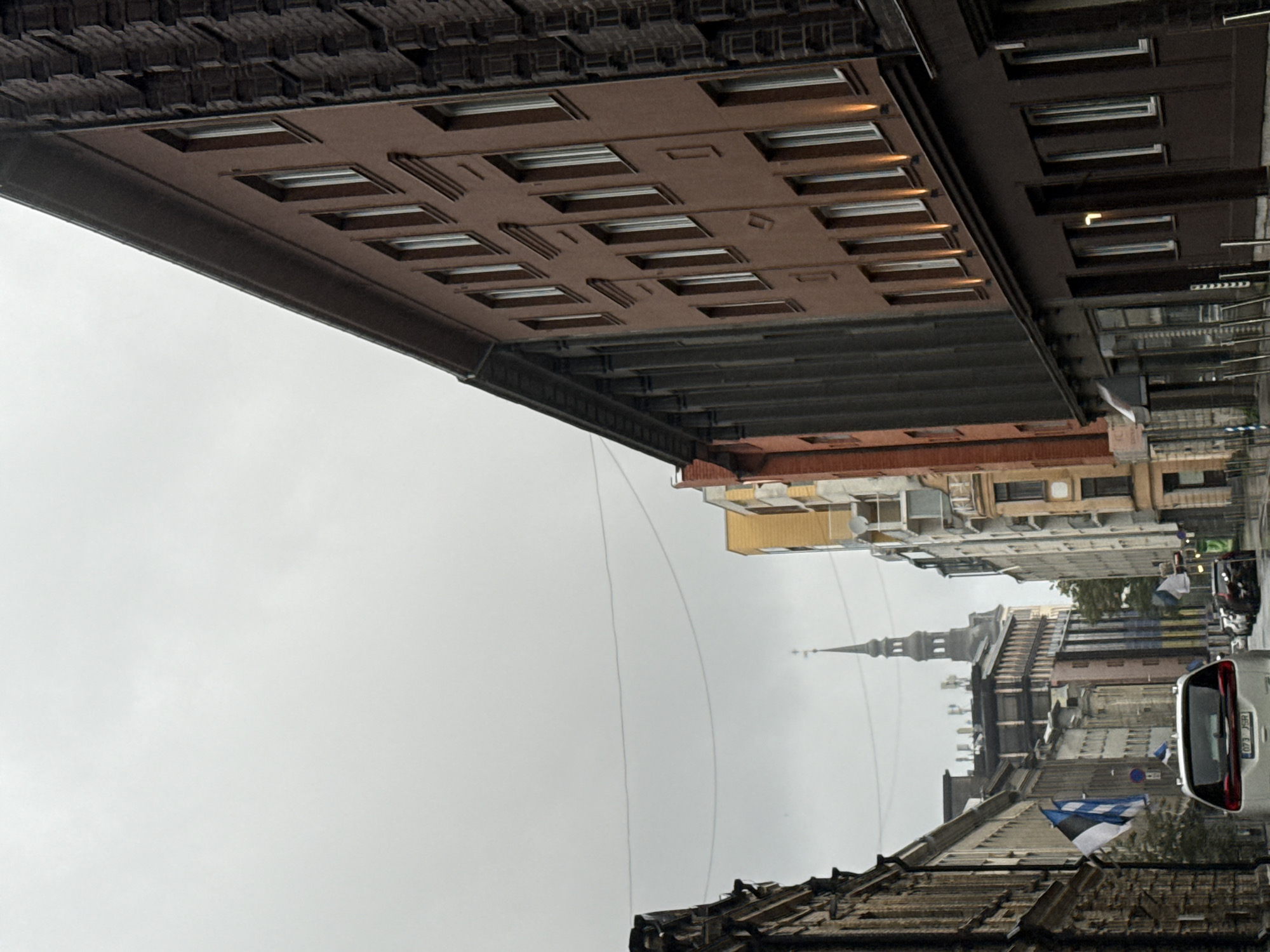 The image depicts a view down a city street in Tallinn, Estonia, likely during overcast weather. The perspective is from a vehicle looking forward. On the right side of the street, a multi-story building with a dark brown facade and numerous windows is prominent. To the left of this building, another building with a lighter brown facade and decorative elements is visible. Further down the street, a tall, spire-topped tower, possibly a church steeple, rises above other buildings. Several Estonian flags, which are blue, black, and white, are displayed on poles along the street. Various cars are parked or driving on the street, including a white SUV in the foreground with a license plate reading "073 JSR". The buildings lining the street exhibit a mix of architectural styles and colors. Overhead, power lines are visible against the cloudy sky. The scene conveys a sense of urban street life with a historical backdrop.
