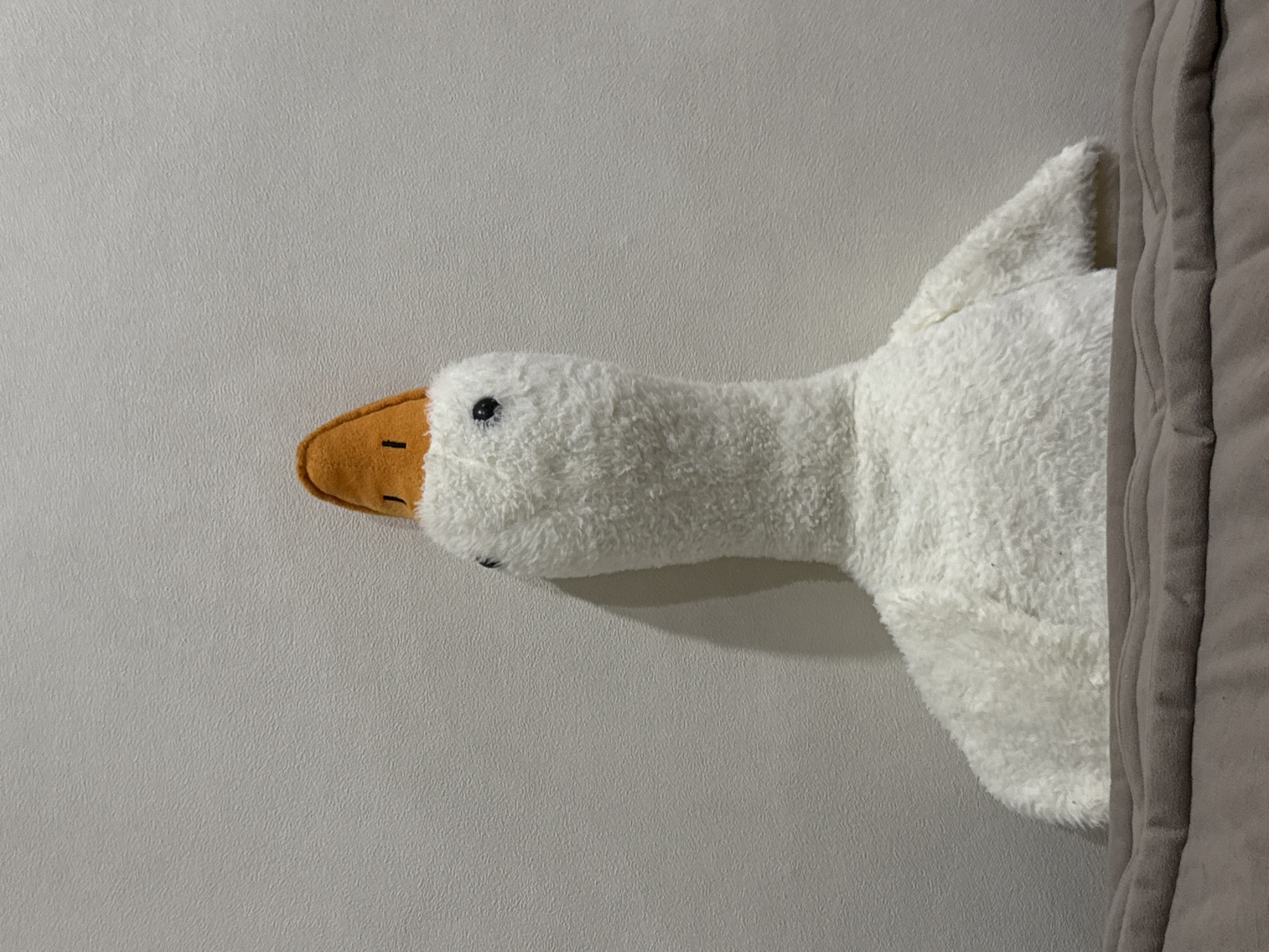 This image displays a white plush goose toy, characterized by its fluffy texture, small black eyes, and an orange, pointed fabric element resembling a hat positioned on its head. The goose is situated against a plain, light beige or off-white wall with a subtle, irregular textured pattern. Faint shadows are cast on the wall to the right of the goose, suggesting a light source from the left. A soft, brown or taupe-colored fabric surface, exhibiting gentle undulations, occupies the lower foreground of the image, partially obscuring the base of the plush toy. The scene is static, devoid of human presence or any discernible action, and is located within an indoor environment in the city of Chaikovsky, Russia.