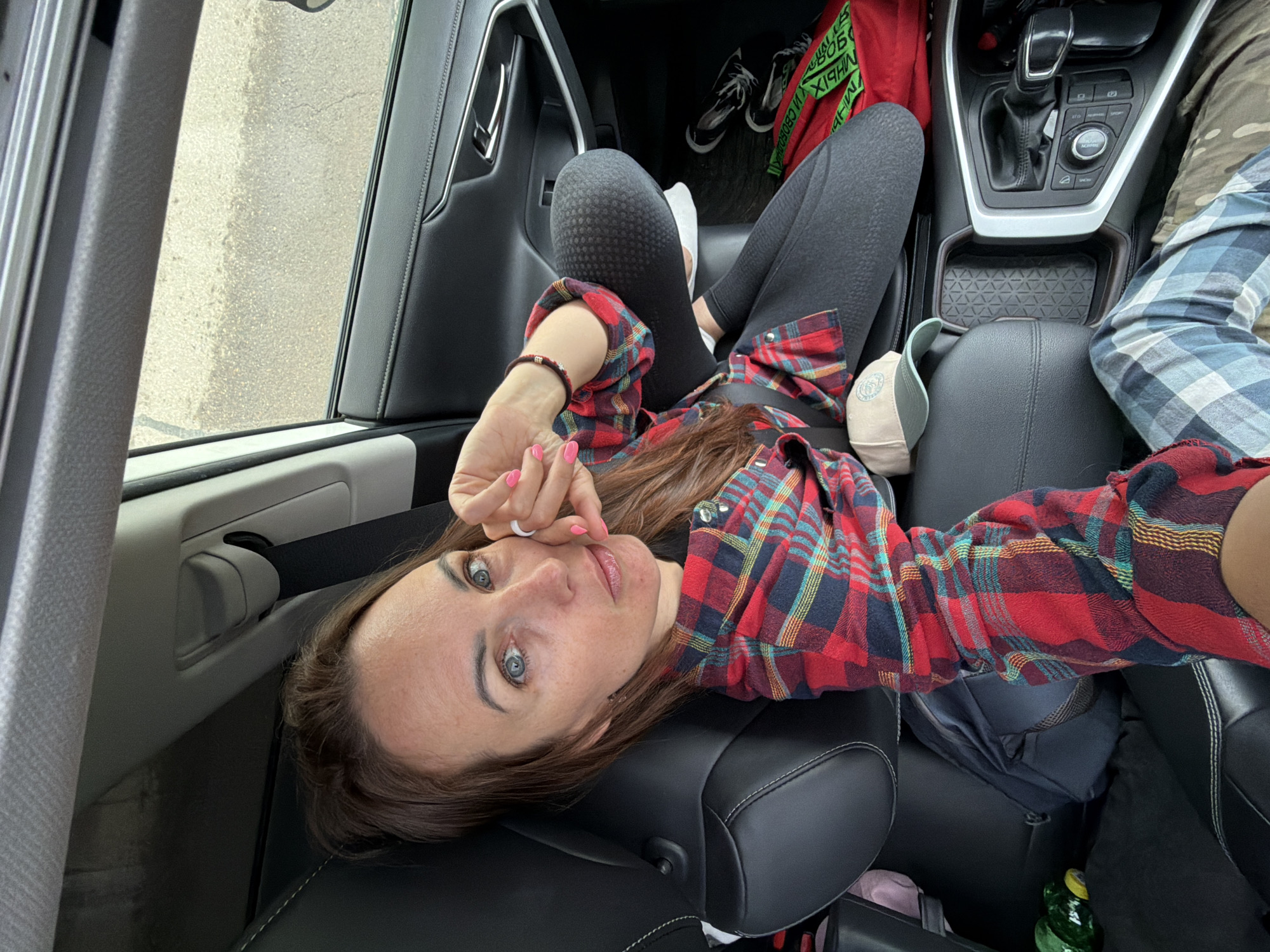 A woman with long brown hair and blue eyes is sitting in the passenger seat of a car. She is wearing a red and black plaid shirt over a black top, black leggings with a textured pattern, and plaid pants. She has bright pink nail polish on her fingernails. She is resting her head on her hand and looking at the camera. The interior of the car is visible, including the black leather seats, the dashboard, and the center console. There are also some items in the car, such as a backpack, a hat, and a red bag. The car appears to be parked on a paved surface, possibly a street or parking lot. The background is blurred, but it appears to be an outdoor setting. There is no specific information to identify the location in the city of Кирга, Russia.