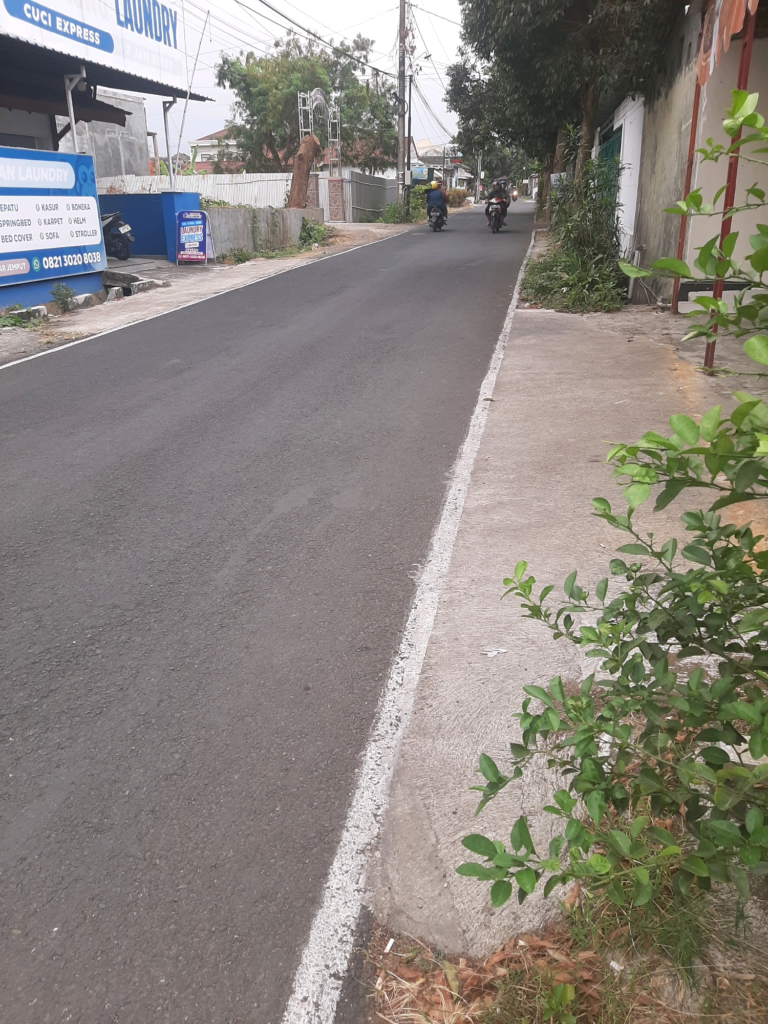 In Sumampir, Indonesia, a street scene unfolds with a road in the foreground and a laundry business on the left. The road, paved with asphalt, features a white painted line along its right edge. A sidewalk runs parallel to the road, constructed from concrete, and is partially obscured by a leafy green plant. On the left, the "LAUNDRY EXPRESS" business is visible with its signage. A blue sign lists services including "KEPATU," "KASUR," "BONEKA," "SPRINGBED," "KARPET," "HELM," "BED COVER," "SOFA," and "STROLLER." A phone number, "0821 3020 8038," is also displayed. Two motorcycles are seen on the road, one with a rider wearing a yellow helmet, heading away from the camera. In the background, trees and buildings are present, and an arched structure with cut trees is visible behind a fence.