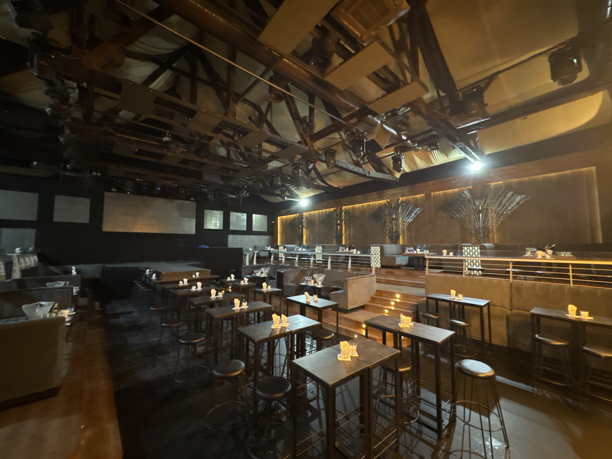 The image shows the interior of a nightclub or lounge in Tsarevo, Bulgaria. The space is dimly lit with a focus on ambient lighting and stage lighting fixtures suspended from the exposed industrial-style ceiling. The room is furnished with numerous high-top tables and stools, arranged in rows throughout the main floor. Some tables are set with small decorative items, possibly candles or flowers, and glassware. In the background, there are elevated seating areas with plush booths and tables, separated by railings. The flooring appears to be dark and possibly reflective, adding to the atmospheric lighting. The overall aesthetic suggests a modern and upscale entertainment venue. There are no people visible in the image.