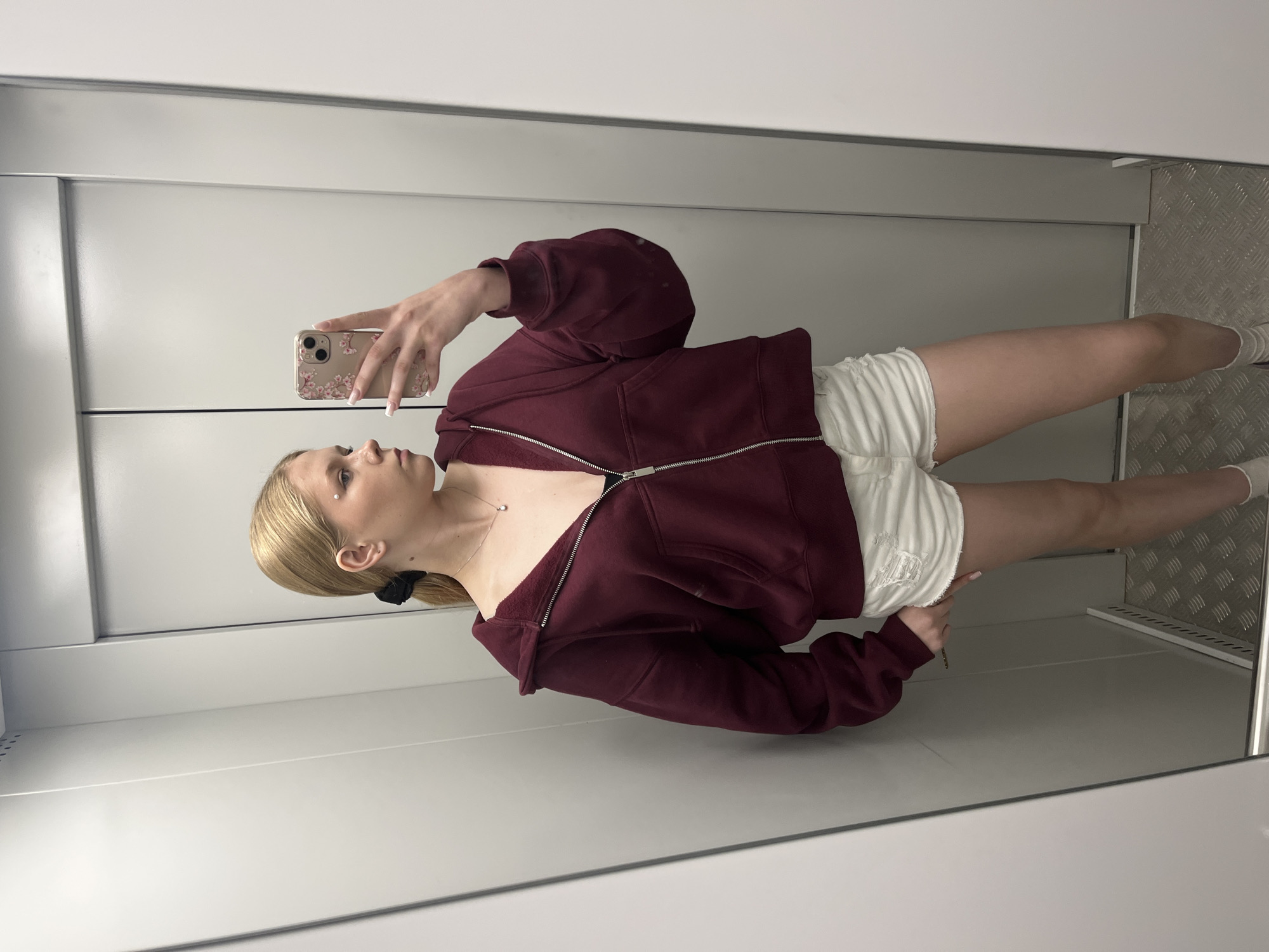 The media file shows a young woman taking a selfie in a reflective surface, likely an elevator or changing room. She has blonde hair styled in a ponytail and is wearing a maroon zip-up hoodie and white shorts. The hoodie is partially unzipped, revealing a delicate necklace. Her nails are painted a light pink. The background is a plain, modern interior with gray walls and a metallic textured floor. The overall impression is casual and personal, with no specific event or time of day indicated. There is no visible text in the image.