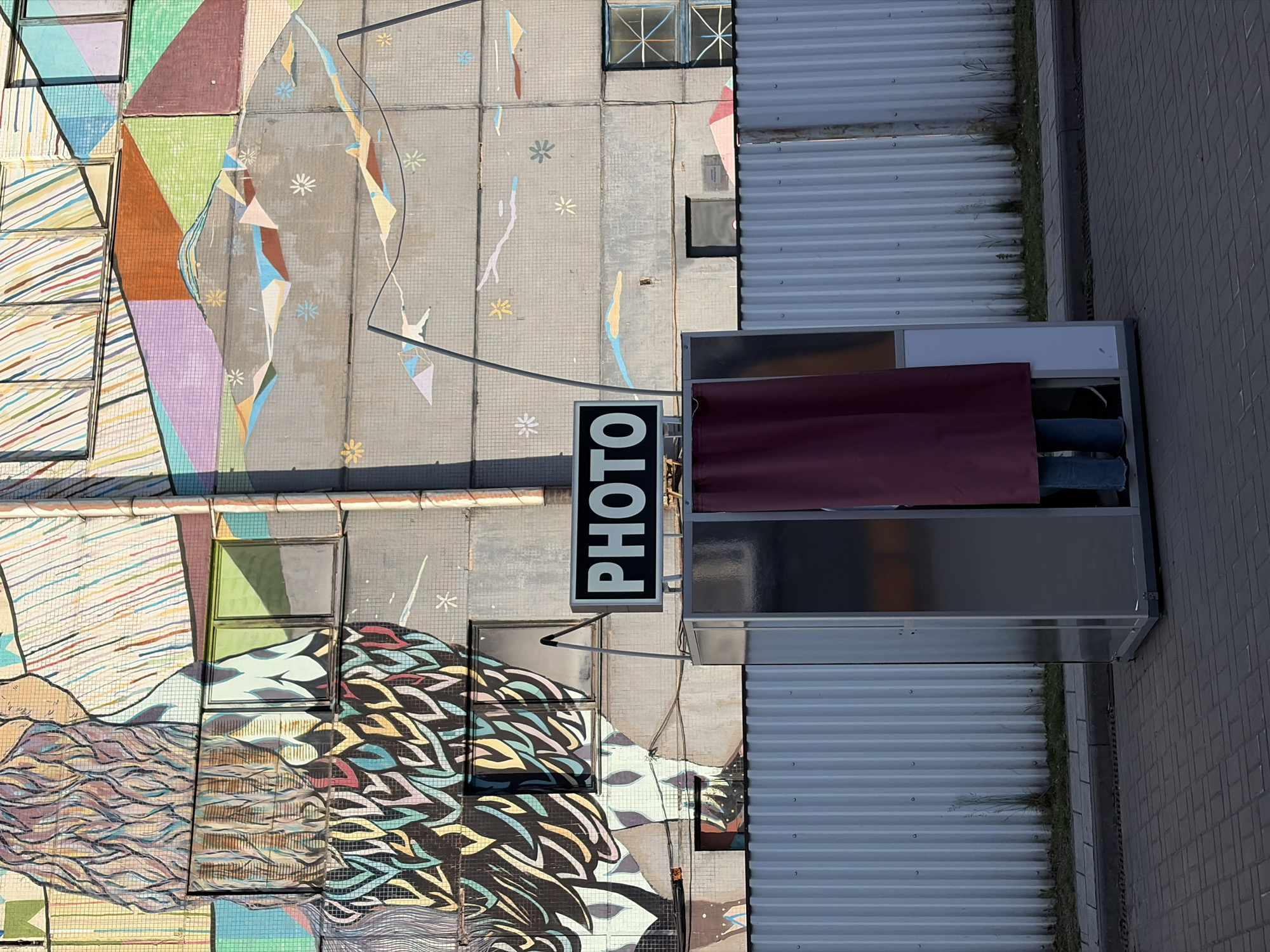 The image shows an overhead view of a photo booth situated adjacent to a building with elaborate murals in Minsk, Belarus. The murals display a variety of designs, including geometric shapes, human forms, and abstract patterns. The photo booth is silver with a maroon curtain visible inside. A sign reading "PHOTO" hangs perpendicular to the booth. Adjacent to the photo booth is a corrugated metal surface with some vegetation growing along the edges. The ground is paved with gray bricks. The photograph shows sunlight, suggesting it was taken during daylight hours.