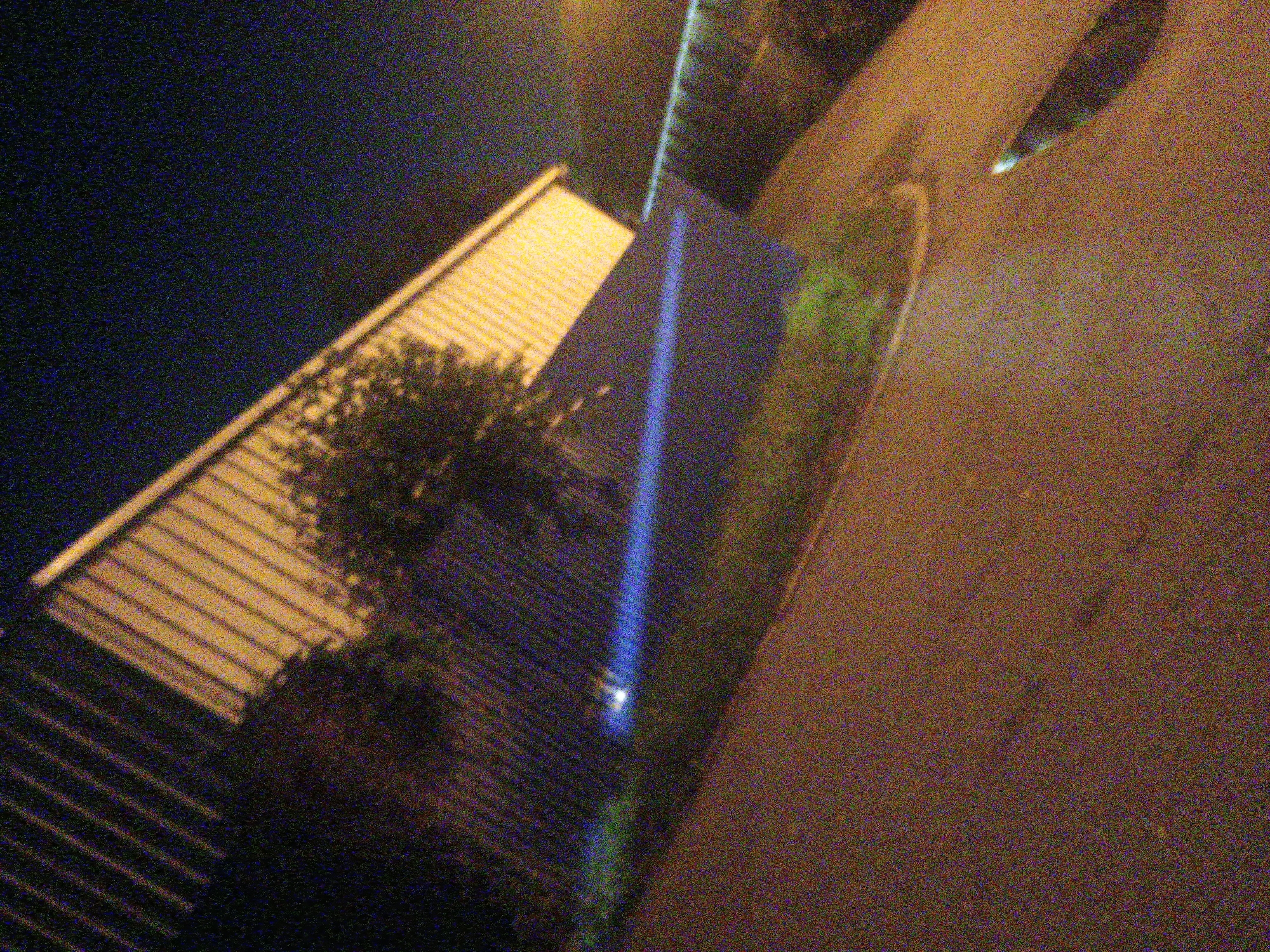 This nighttime image, captured from an elevated, angled perspective, depicts a scene in Kroměříž, Czechia, heavily impacted by digital noise due to low-light conditions.
On the left side of the frame, a building with a light-colored, possibly corrugated or ribbed roof is partially visible. A tree or dense shrubbery stands in front of this building. A prominent, intense beam of bright blue light slices vertically through the center of the image, appearing to originate from a source in the lower portion of the frame and extending upwards. This light illuminates a section of a darker, vertical surface, possibly a wall or another structure, before fading into the dark background. To the right, a broad, textured area, likely a road or paved ground, is discernible.
No specific activities or individuals are visible. The overall impression is that of an outdoor urban or suburban setting at night, with the striking blue light suggesting a deliberate lighting display, event illumination, or perhaps a spotlight. No visible text is present.