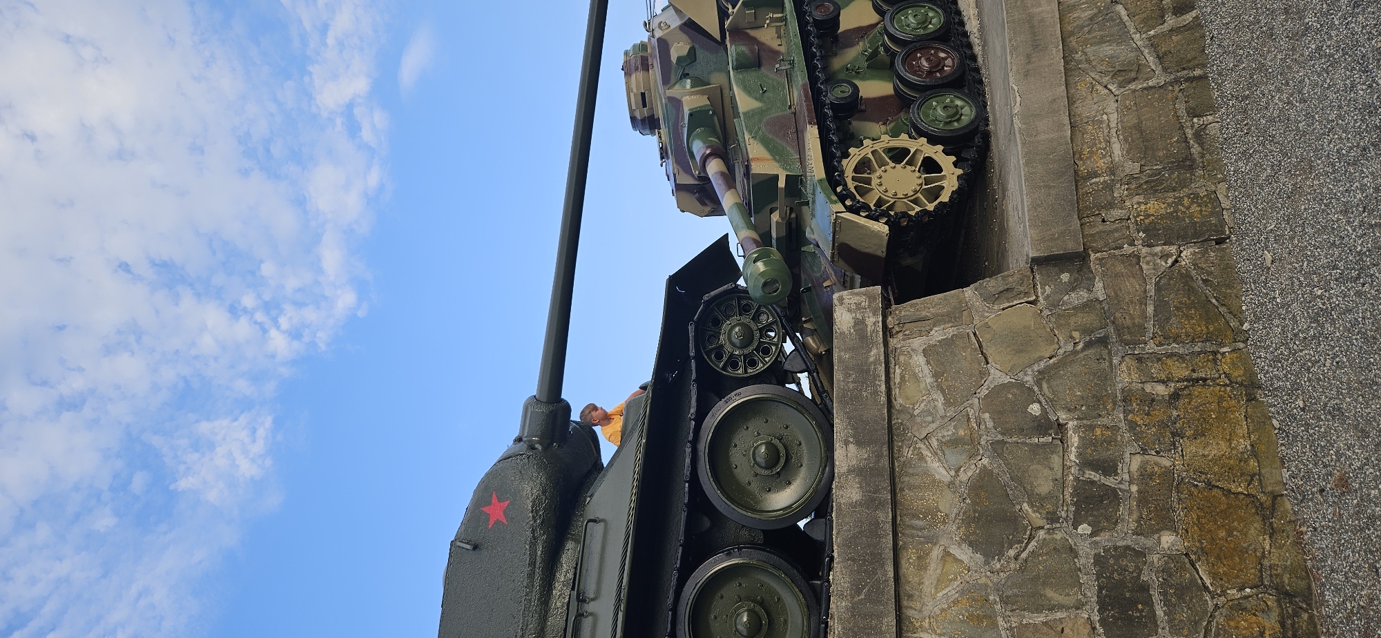 In Svidník, Slovakia, two military tanks are displayed on a stone embankment. The tank on the left, painted dark green with a red star, has a young boy in an orange shirt sitting on its hull. The tank on the right is camouflaged in greens and browns. The sky above is blue with scattered white clouds. The embankment is constructed of large, irregularly shaped stones. The foreground consists of gravel.