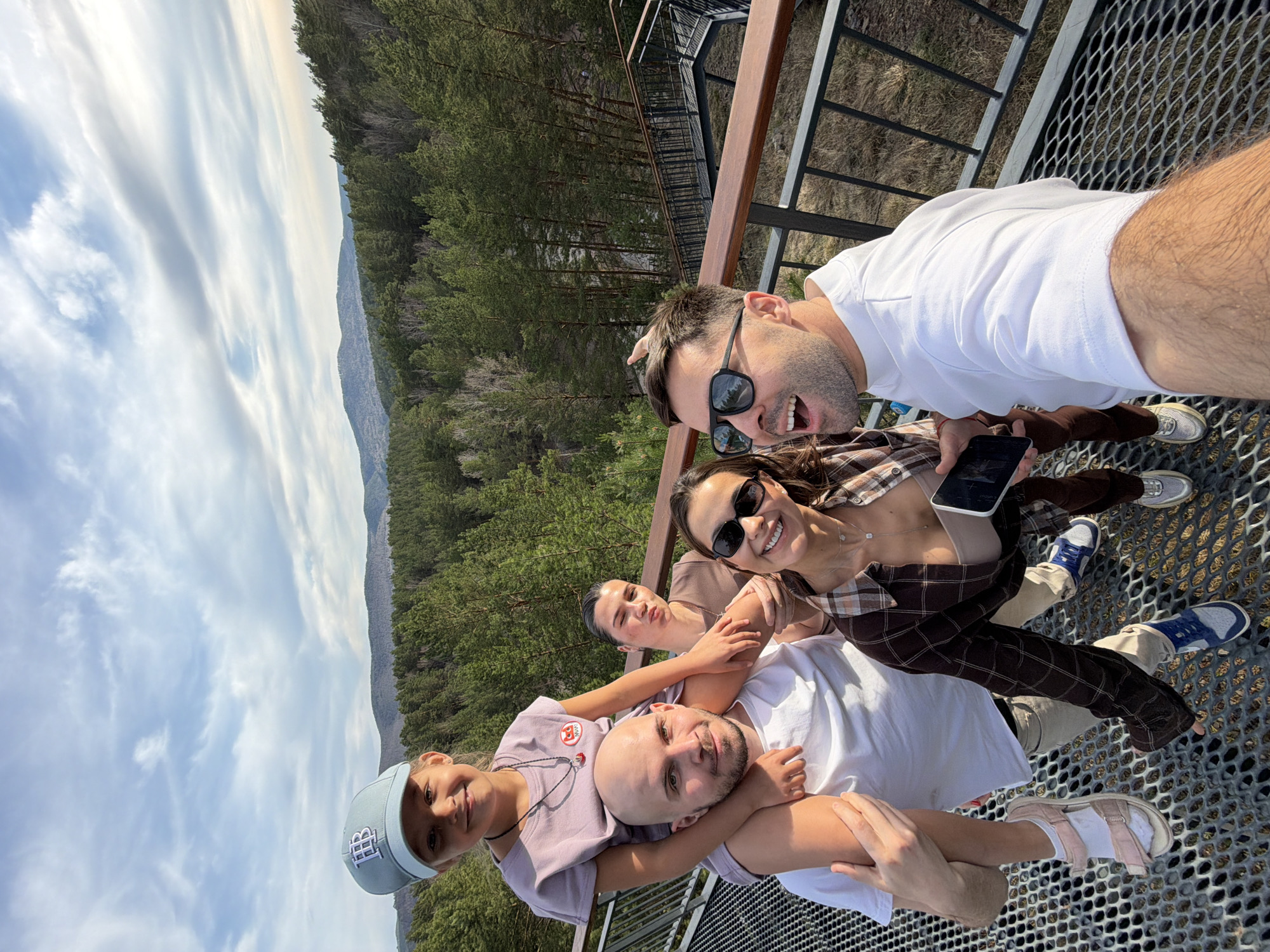 The image shows a group of five people taking a selfie on a metal grid platform with a railing. In the background is a forest and mountains under a sky with scattered clouds. The individuals are arranged in a pile, with one person on the bottom, another lying on top, and the rest clustered around the top. The person taking the selfie is on the top right and is wearing sunglasses and a white shirt. Another person is holding a smartphone. The location is likely the Tsar Fish viewing platform in Krasnoyarsk, Russia.