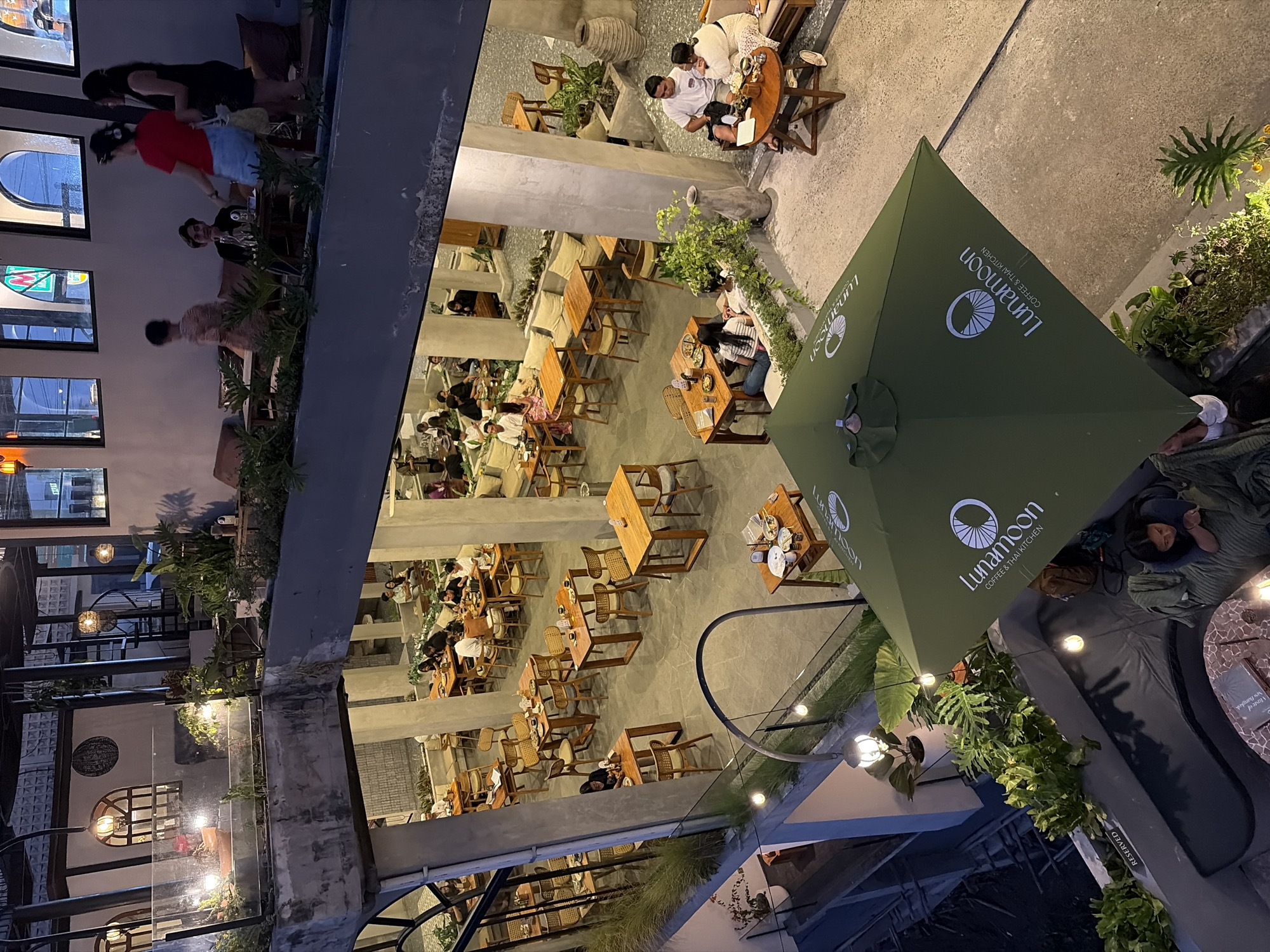 This image captures an outdoor seating area of a cafe at night, with people dining and socializing. The scene is illuminated by ambient lighting, suggesting a warm and inviting evening atmosphere. The cafe features an array of wooden tables and chairs, many of which are occupied by patrons. Lush greenery is interspersed throughout the space, adding a natural element to the urban setting. A large, green umbrella, prominently displaying the text "Lunamoon COFFEE & THAI KITCHEN," provides shade and branding for the establishment. The overall mood appears relaxed and convivial, as people engage in conversations and enjoy their meals. The presence of lanterns and string lights further enhances the nighttime ambiance. The visible text "RESERVED" suggests specific areas or tables have been pre-booked. The location context of Kintamani, Indonesia, implies a tropical setting, which is consistent with the lush vegetation.