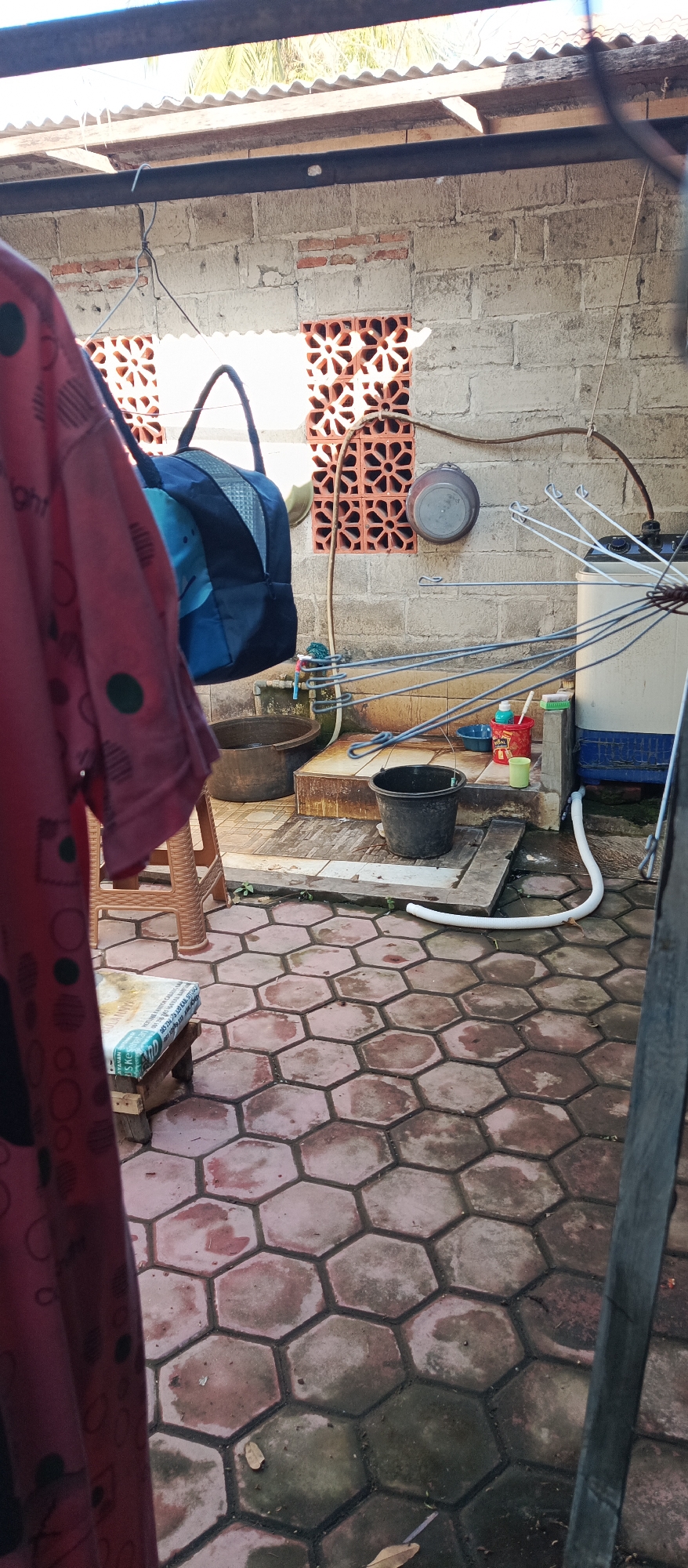 The image depicts a back area of a house, likely a laundry or utility space, in Arjasa, Indonesia. The foreground is dominated by a red garment with a black circular pattern, hanging from a clothesline or hook, partially obscuring a woven plastic chair and a wooden stool with a cushioned top. The ground is paved with hexagonal red and gray tiles, some of which are wet and discolored, with a few dry leaves scattered about.
In the mid-ground, a grey brick wall serves as the backdrop. Two decorative orange brick vents with star-shaped patterns are visible in the wall. A blue and black duffel bag hangs from a hook to the left of the vents. Below the vents, a slightly raised concrete platform holds several items: two large dark-colored basins, a black bucket, a blue washbasin, a red plastic container with cleaning supplies and a brush, and a green cup. A white washing machine with a blue base is situated to the right of the platform, connected to a white corrugated hose that snakes across the tiled ground. A drying rack with numerous wire hangers is attached to the washing machine. A round metal pot is hanging on the wall above the platform.
The background shows a corrugated metal roof supported by dark metal beams, with glimpses of palm fronds through the gaps, indicating a tropical environment. The overall impression is of a functional, somewhat cluttered outdoor living or working space.
