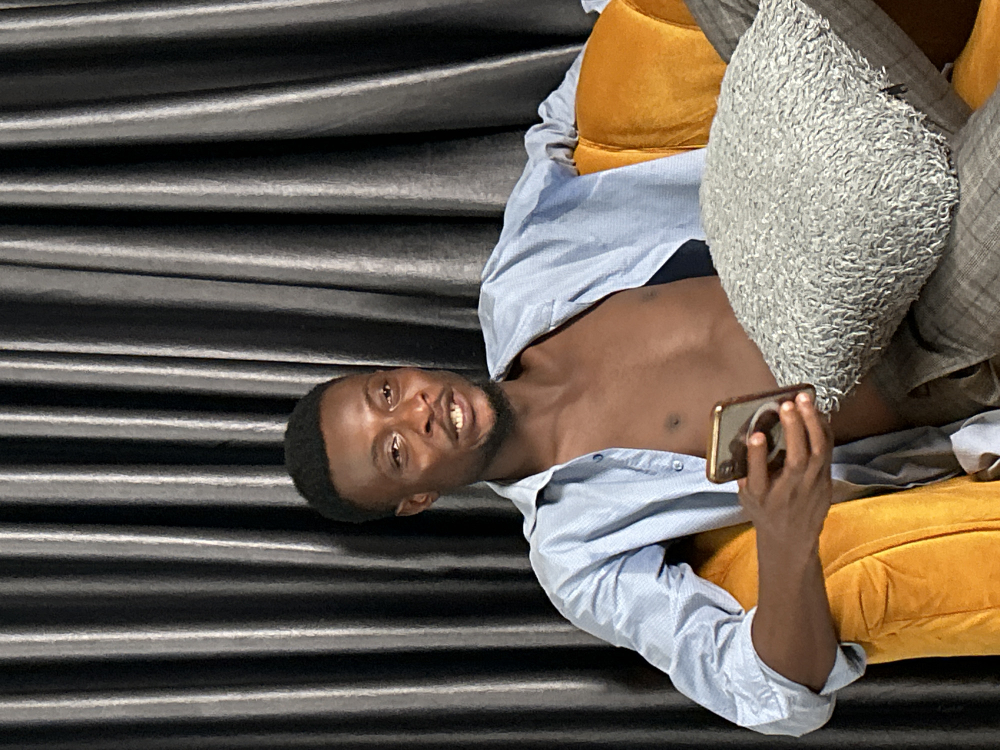 A man with dark skin, a short beard, and a blue button-down shirt, partially unbuttoned, is reclining on a yellow velvet couch with a gray textured pillow, holding a gold-colored smartphone in his right hand, appearing to take a photo or video. The background is a dark gray, textured material resembling drapes or a curtain. The location appears to be indoors, but the city and specific location are Unknown.