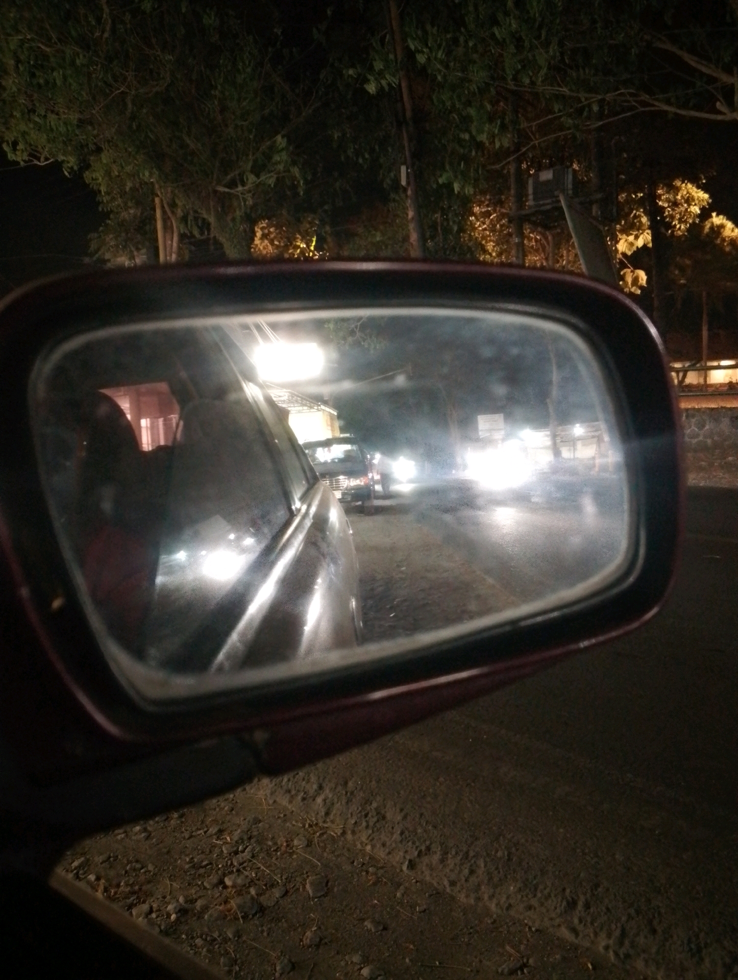 This is a nighttime photograph taken from inside a car, looking into the side mirror. The mirror reflects a road with traffic, trees, and buildings. The scene appears to be a street in Ngebra, Indonesia.
The mirror shows other vehicles on the road, their headlights creating bright streaks of light. In the reflection, there are also some buildings and trees visible. The overall impression is of a busy street at night, possibly with a lot of traffic. The photograph is slightly blurry, which is common for nighttime shots taken with a smartphone.
The location is Ngebra, Indonesia, a city known for its bustling streets and vibrant nightlife. The photograph captures the essence of this lively atmosphere.