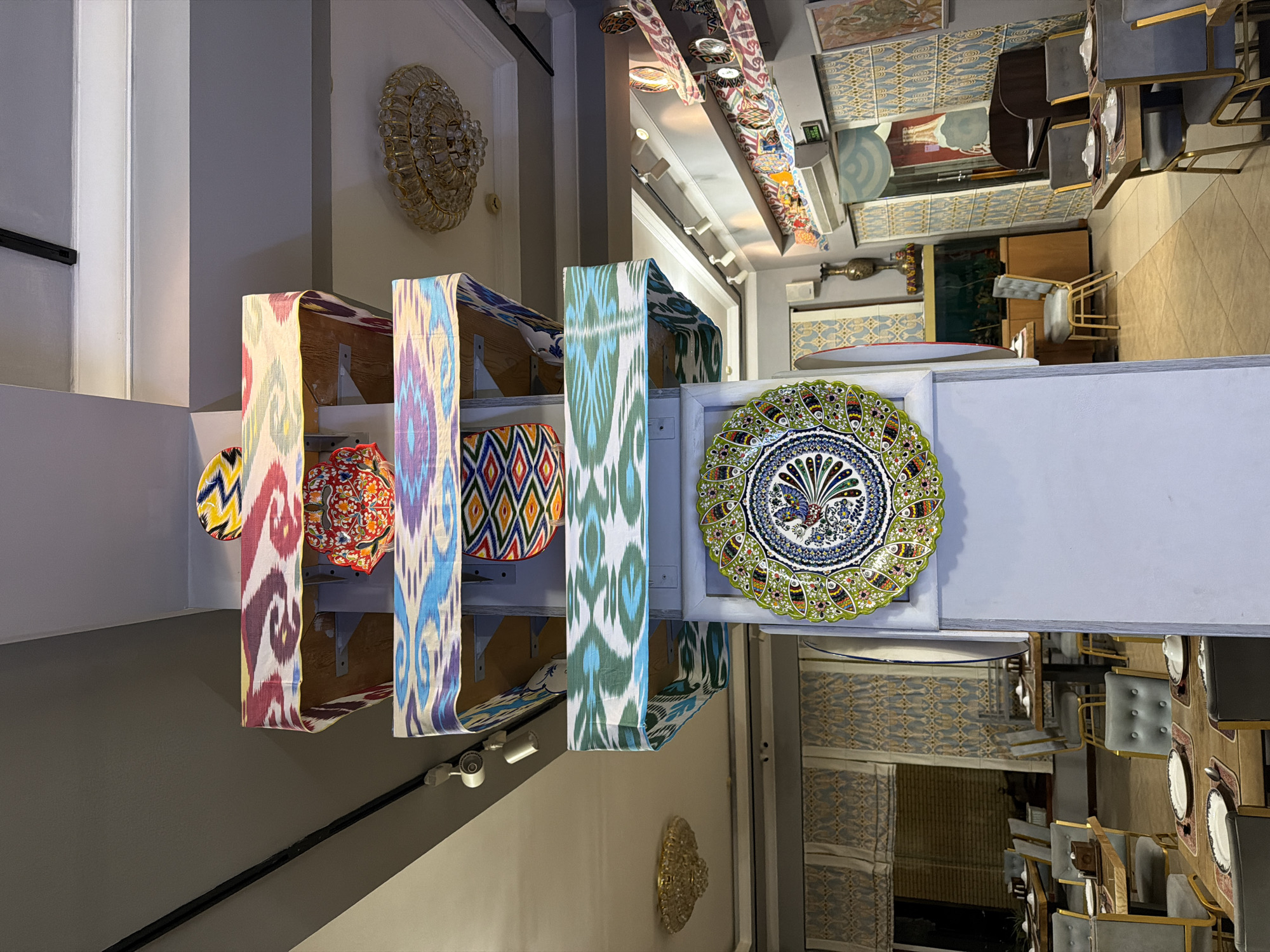 The image depicts an indoor scene, likely a shop or a market stall, with an emphasis on decorative textiles and ceramics. In the foreground, several pieces of vibrantly patterned fabric hang vertically, showcasing intricate ikat designs in shades of red, pink, purple, blue, green, and yellow. Below these, a collection of round, decorative plates is displayed, one with a striking yellow and black geometric pattern, another with a red floral motif, and a large, central plate with a detailed blue and green peacock or bird design.
Behind the textiles and plates, the setting appears to be a retail space. To the left, a portion of a white wall is visible, and on it hangs a large, ornate, circular wall decoration that resembles a chandelier or a sunburst. Further back, a doorway or an opening leads to another section of the shop, where more textiles and decorative items are displayed on shelves and hanging from the ceiling. The overall impression is one of a colorful and culturally rich display of artisanal crafts.
In the background, glimpses of a dining area can be seen, with tables and chairs, suggesting that this might be a shop selling homeware or that it's part of a larger establishment with a dining space. The lighting is artificial, creating a warm and inviting atmosphere. No people are visible in the frame, focusing the attention solely on the objects and the environment. The style of the textiles and ceramics suggests a possible Central Asian origin, consistent with the location context of Chinarkent, Uzbekistan.