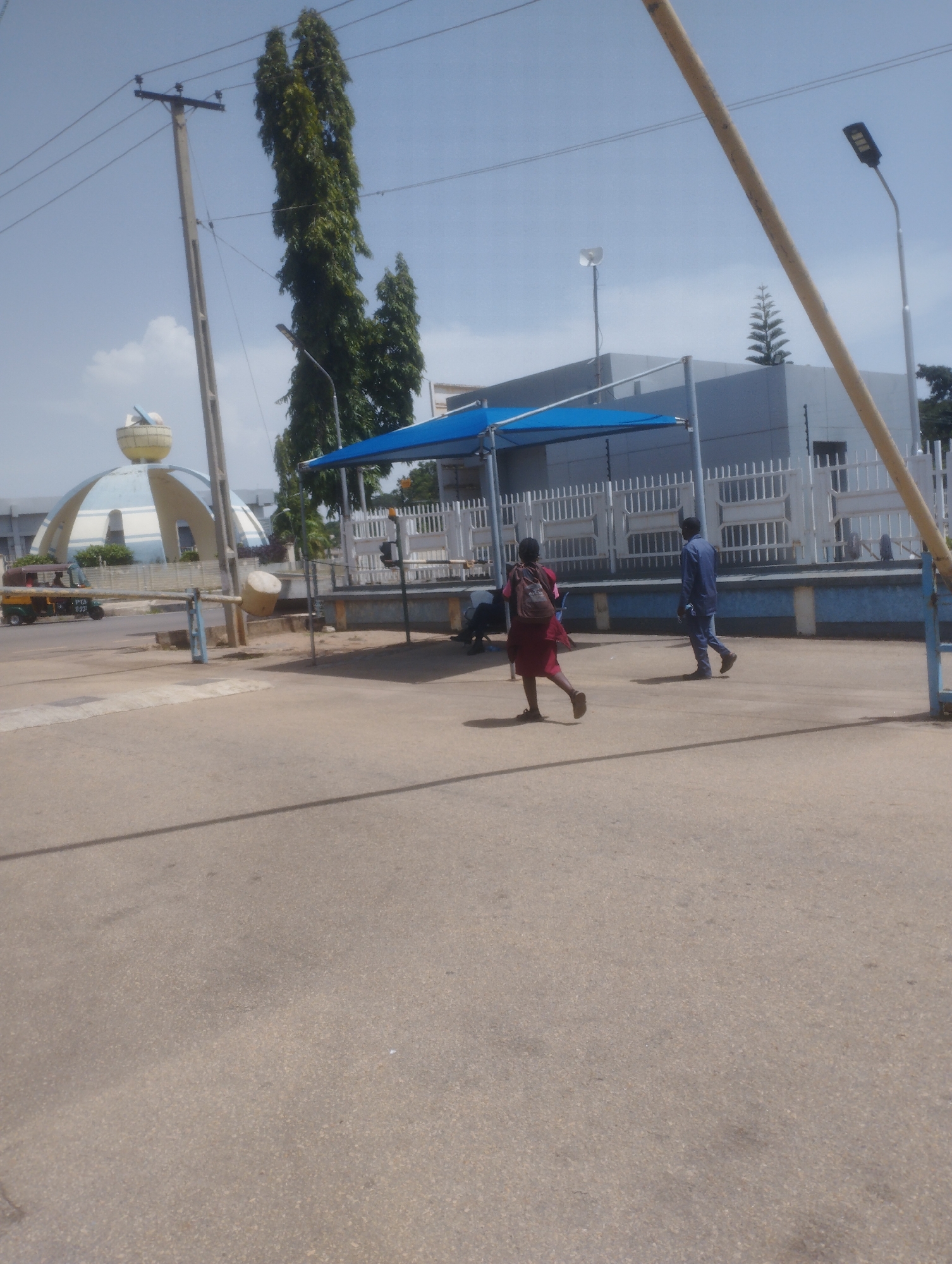 In this sunny daytime scene in Jos, Nigeria, two individuals are walking on a paved area. One, a person in a maroon dress and backpack, is running ahead. Behind them, a person in a blue shirt and pants walks away from the camera.
The background features a white fence and a modern building with a blue awning. In the distance, a unique domed structure with a rounded top and curved sides, reminiscent of a large orb or a stylized dome, is visible. A utility pole with electrical wires stands prominently on the left side of the image. A three-wheeled auto-rickshaw, commonly known as a "Keke Napep" in Nigeria, is seen driving past on the left. The sky is clear with a few wispy clouds. The overall atmosphere appears to be that of a normal day, with people going about their activities in an urban setting.