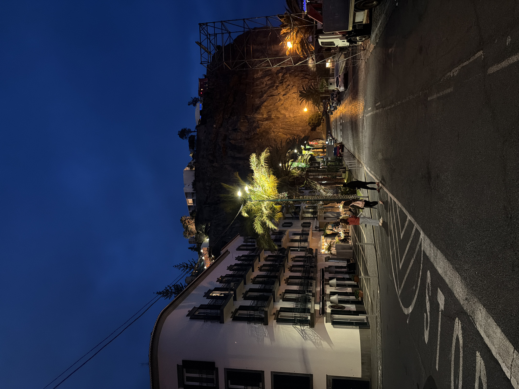 The image is a vertical, nighttime shot of a street scene in Ponta do Sol, Portugal. On the left, a multi-story white building with numerous black-shuttered windows is lit up, with a tall palm tree illuminated in front. People are seen along the street, some appearing to be dining outdoors. Behind the building and across the street, there's a rocky cliff illuminated with lights and what appears to be a scaffolding structure on top. A road with a painted stop sign runs along the right side, and several parked cars are visible further down the street. The sky is a dark, clear blue.