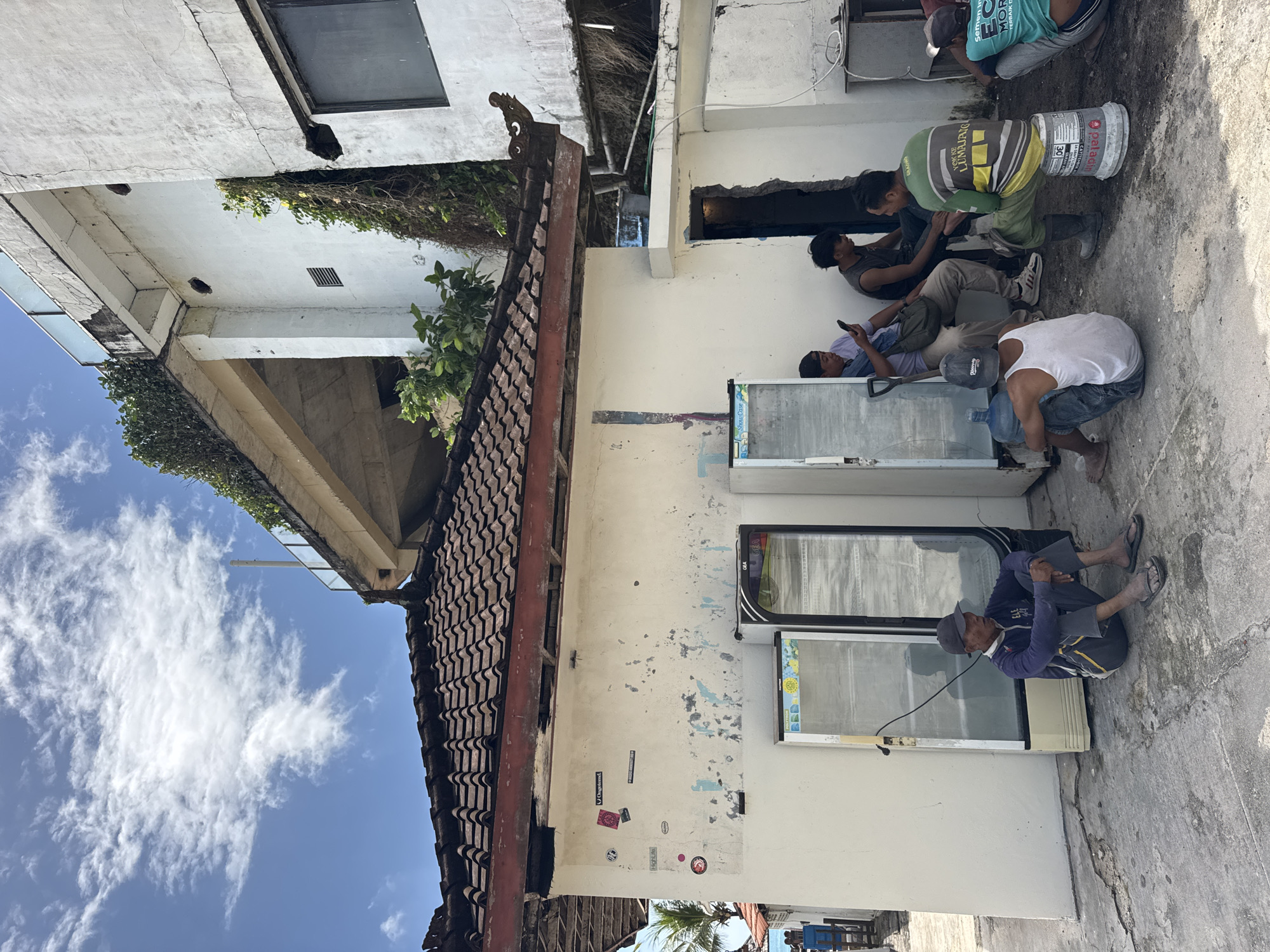The image shows an outdoor scene in Kuta Selatan, Indonesia. Several construction workers are resting near a white building. The building has two large windows or display cases and peeling paint. Stickers adorn the wall. A partially visible traditional Balinese roof borders the structure. One worker is using his phone, one is leaning back, and another is crouching near a water bottle. A worker in a green shirt uses a bucket for leverage. The sky above the building is blue with scattered white clouds. The ground is concrete.