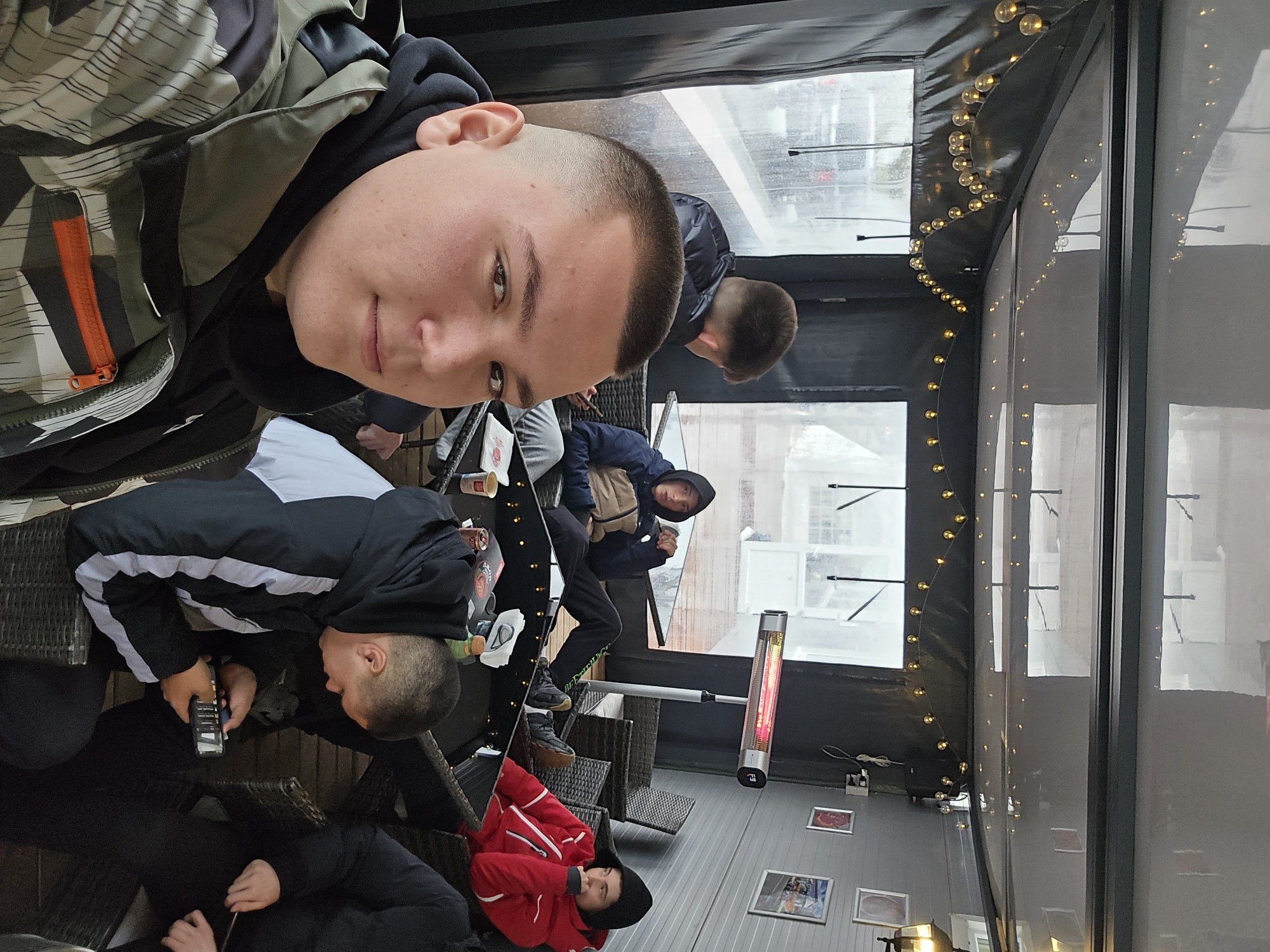 The image shows a group of people in what appears to be an outdoor cafe or enclosed patio area in Samokov, Bulgaria. The cafe has windows and a black covering with string lights. It looks like it's raining or snowing outside. There are several people visible: One person is taking the photo. Another person is looking at a phone. Another person is wearing a red jacket. Another person is standing. There is also a heater in the space, and several framed photographs are placed near the floor.