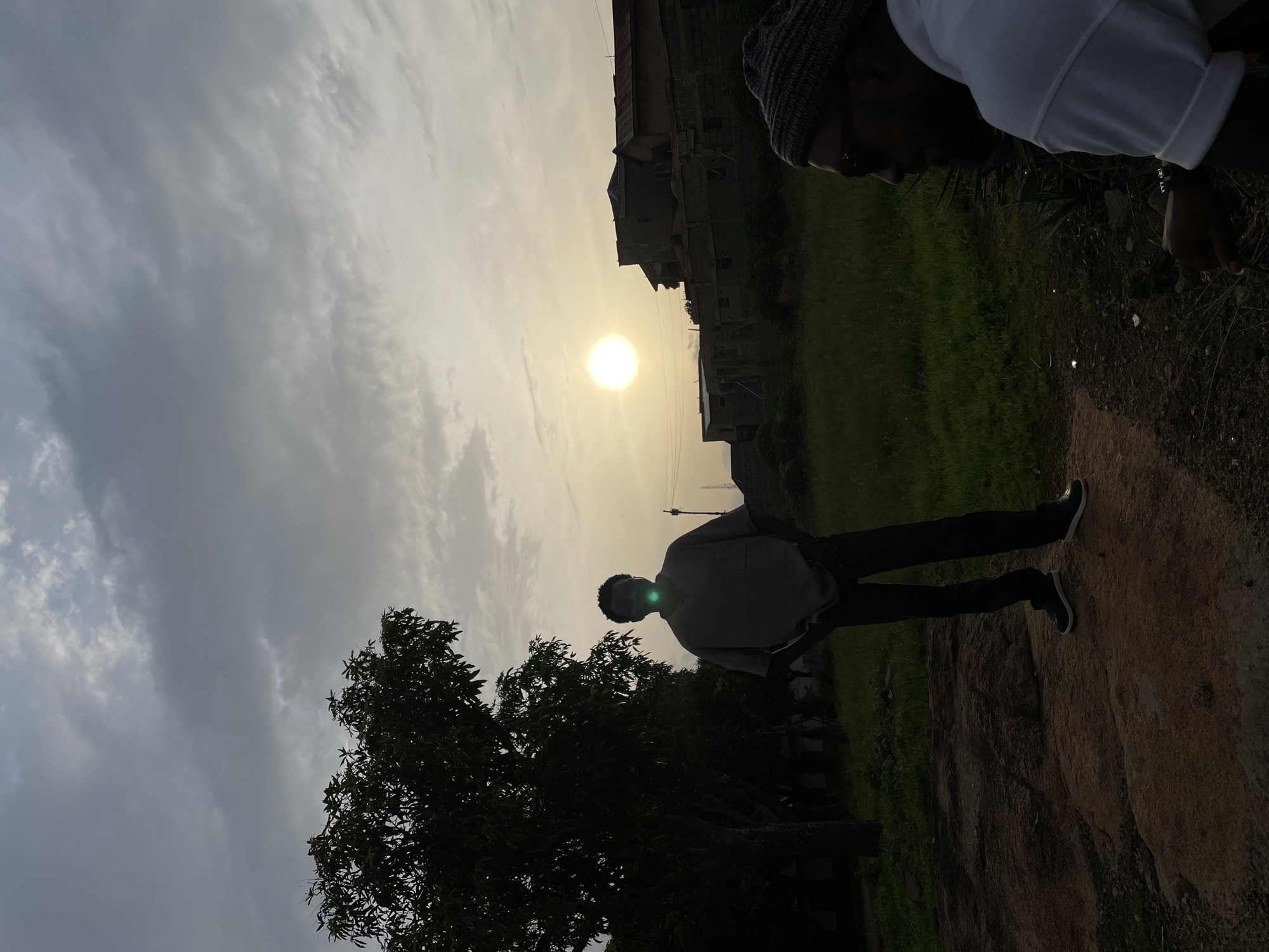 The image depicts an outdoor scene at sunset in Jos, Nigeria. A person is standing in the foreground, their silhouette outlined against the bright sun and cloudy sky. To the left of the person is a large tree, also silhouetted. Behind the person, the ground slopes upwards and is covered with green grass and some patches of bare earth. A building is visible in the background, partially obscured by the sun's glare. Another person is partially visible in the upper right corner of the image. The sky is filled with clouds.