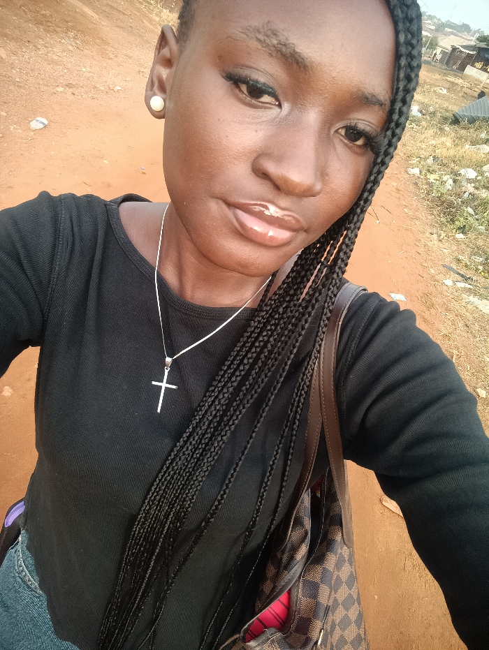 A woman poses for a selfie outdoors. She has dark skin, braided hair, and wears a black long-sleeved shirt. A silver necklace with a cross pendant adorns her neck. She has on a pearl earring. A checkered brown bag hangs off her shoulder. The background reveals a dry, earthy landscape with some distant buildings. Her lips are glossy.