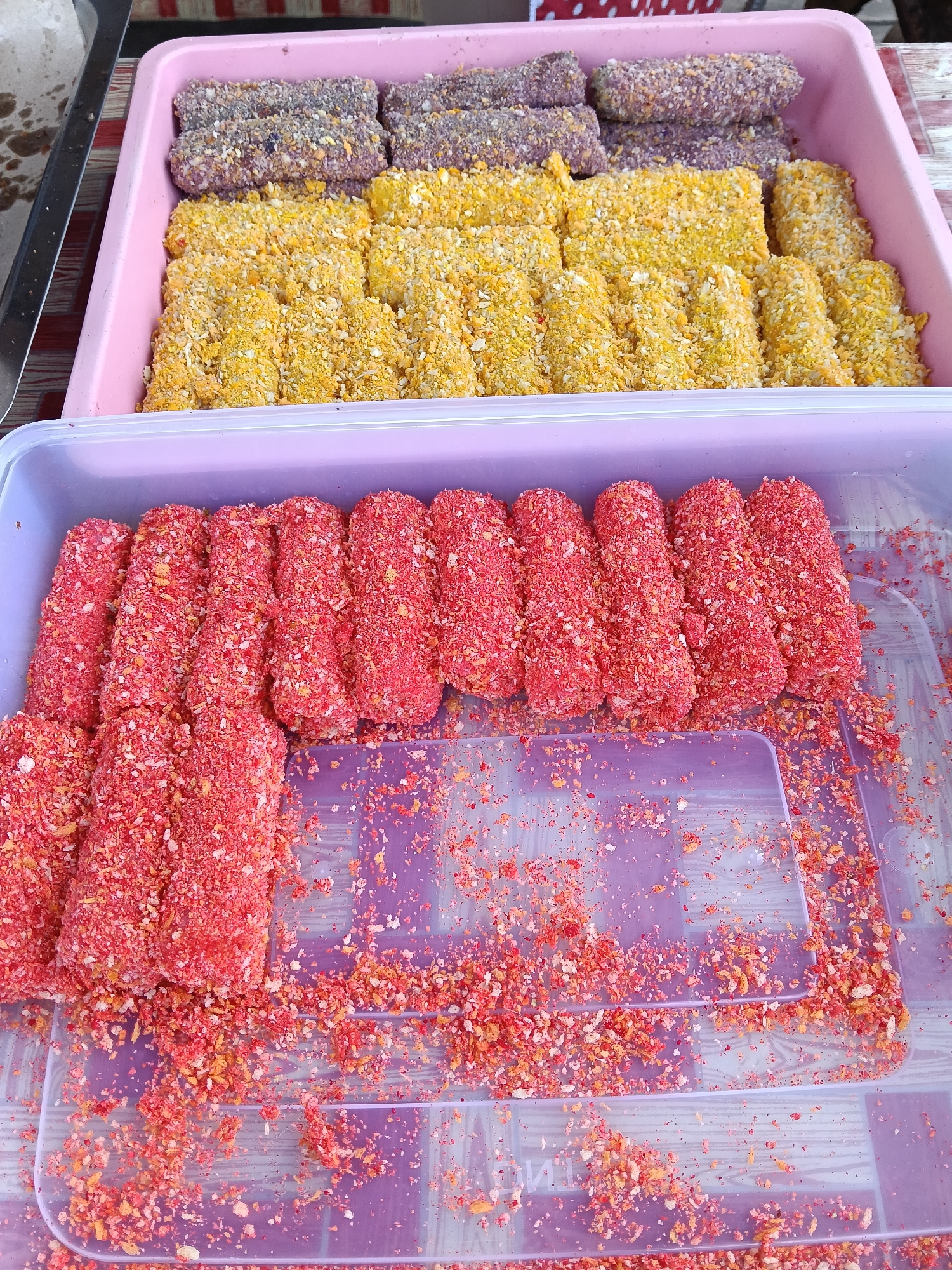 The image shows two rectangular plastic trays containing different types of food. The top tray is pink and holds items coated in yellow and purple granular material. The bottom tray, made of light purple plastic, contains items covered in a bright red granular coating, with loose coating material visible at the bottom of the tray. The image appears to be taken at an elevated angle. Given the location in Lhokseumawe, Indonesia, it is likely these are snacks or small food items sold at a market or roadside stall.