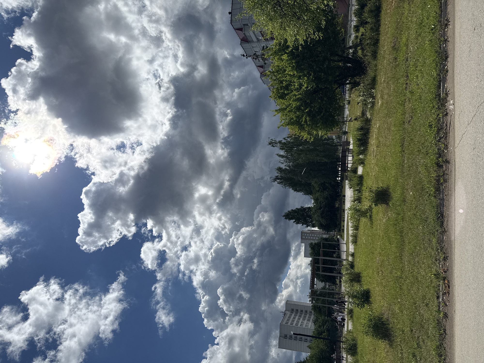 The image depicts a cloudy sky with patches of bright sunlight and a blue sky. Below the sky, there's a grassy area with some trees and buildings in the background. A paved surface runs along the right side of the image, suggesting a sidewalk or road.
The subjects in the image include:
* **Sky:** Dominated by large, puffy white and dark gray clouds, with the sun partially visible and creating a lens flare effect.
* **Vegetation:** Lush green grass covers the ground, and several trees with dense foliage are present.
* **Buildings:** Residential buildings, likely apartment blocks, are visible in the background, characterized by multiple windows and light-colored facades.
* **People:** Several small figures of people are visible in the mid-ground, suggesting activity on a pathway or promenade.
The scene appears to be an outdoor urban or suburban environment. The presence of grass, trees, buildings, and a paved path indicates a park-like area adjacent to residential buildings.
Notable details:
* **Time of Day:** The bright sunlight and shadows suggest it is daytime, likely late morning or afternoon.
* **Weather:** The sky is partly cloudy, with both sunny and overcast conditions apparent.
* **Location Cues:** The architectural style of the buildings and the general environment are consistent with a location in Russia.
There is no visible text in the image. The overall mood is one of an ordinary day, with a dynamic sky adding visual interest to the scene.