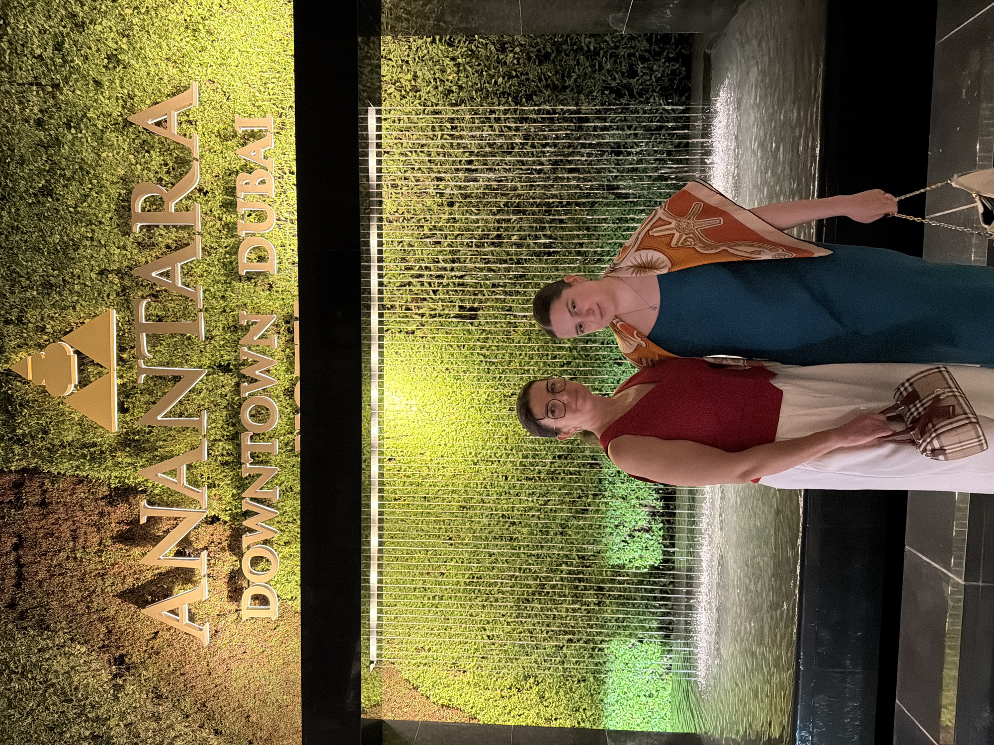 Two women stand in front of a vertical green wall with water cascading down it. Behind them, the words "ANANTARA DOWNTOWN DUBAI" are lit up in gold lettering. The woman on the left wears a red top and white pants, and carries a plaid handbag. The woman on the right wears a teal dress and an orange and white patterned scarf, and carries a light-colored handbag with a chain strap. The setting appears to be an outdoor or semi-outdoor area at night.