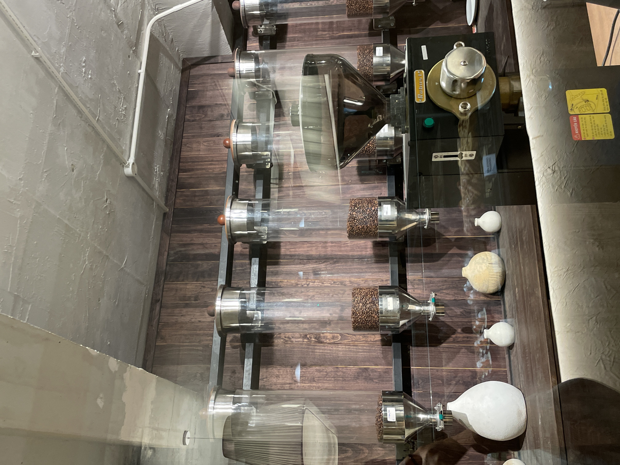 In the city of Da Lat, Vietnam, a commercial coffee grinder is displayed behind a glass partition, likely in a cafe or roastery. The grinder is black with metallic gold accents and features a large hopper filled with coffee beans at the top, which are dispensed into a lower grinding chamber. Several tall, clear cylindrical containers filled with coffee beans are arranged above the grinder on a metal shelf, each with a silver dispenser mechanism at the bottom. The background is a dark, vertically paneled wooden wall. In the foreground, a wooden shelf displays several decorative ceramic vases of varying shapes and sizes. The ceiling is rough concrete with visible electrical conduits. The scene is well-lit, highlighting the textures of the wood, concrete, and ceramic objects.