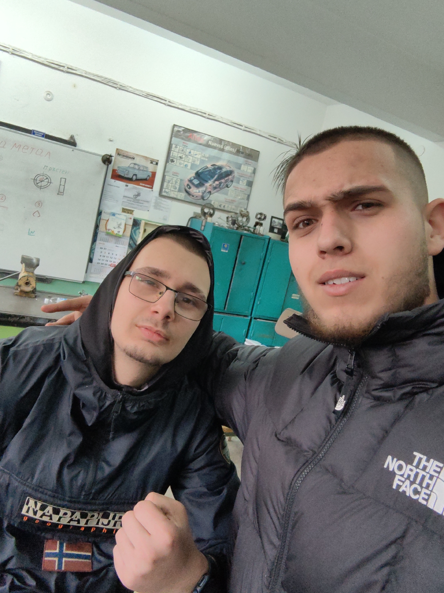 Two young men are posing for a selfie indoors. The man on the right has short, dark hair and a beard and is wearing a black North Face jacket. The man on the left is wearing a black hooded jacket with a Napapijri logo and the Norwegian flag on the front, along with glasses. In the background, there is a whiteboard with the words "метол" and "Пръстен" written on it, along with some diagrams. There is also a poster showing diagrams of cars and car parts. Behind them are light blue metal cabinets. Given the Cyrillic script on the whiteboard, the image could be taken in Sofia, Bulgaria, or another location that uses Cyrillic script.