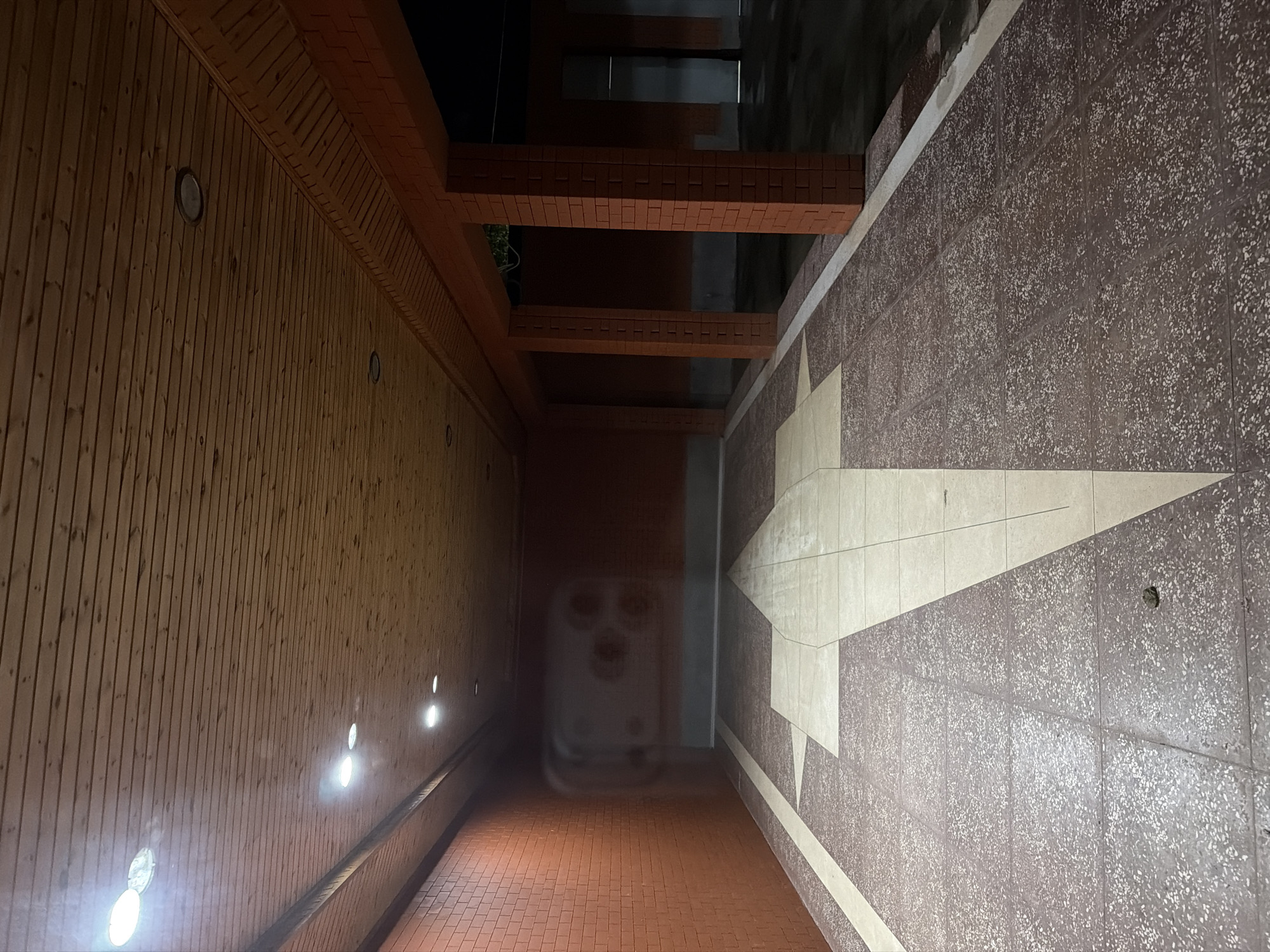The image depicts an outdoor covered walkway or passage with brick walls and columns. The ceiling is made of wooden planks, and recessed lighting illuminates the space. The floor is tiled with a patterned terrazzo material, featuring a large arrow-shaped design in lighter tiles. On the left, a brick wall is visible, and on the right, there are brick columns supporting the roof structure. The walkway leads into a darker, seemingly open area or another building. There are no people or animals visible in the scene. The overall impression is of an architectural space, possibly part of a public building or a protected walkway in Kurchaloy, Russia.