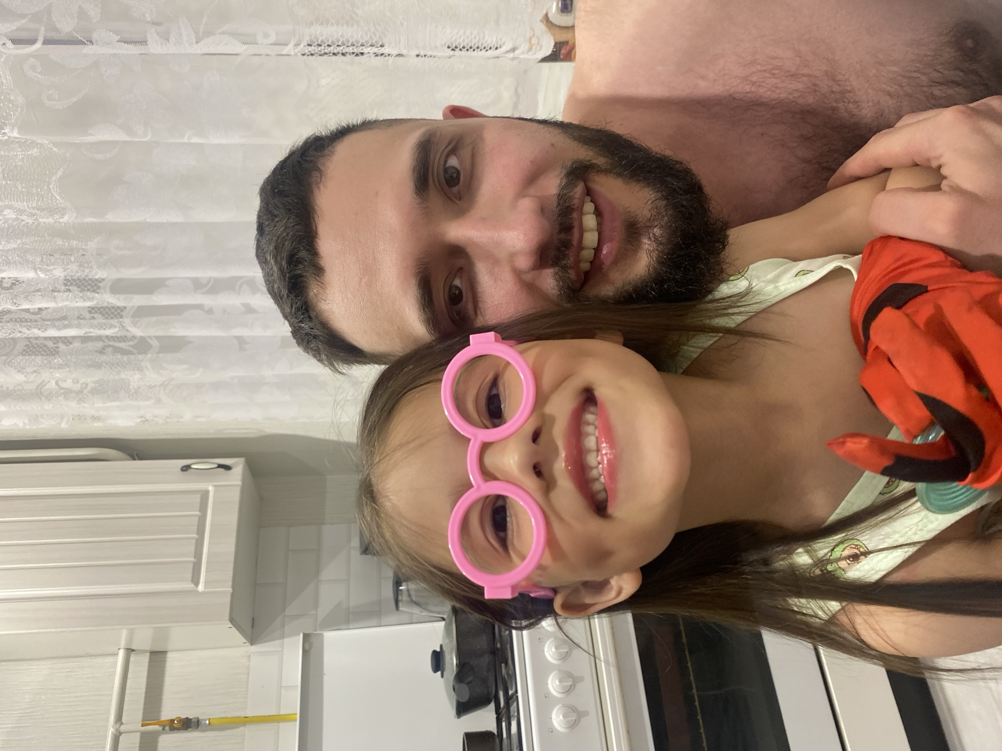 An indoor, close-up photograph captures an adult man and a young girl smiling cheerfully. The image is oriented sideways, with both subjects looking towards the camera.
The man, positioned behind the girl, has a dark beard and dark hair, and is bare-chested, showing a hairy chest. He smiles broadly, revealing his teeth, and his arm is seen gently around the girl.
In the foreground, a young girl with long dark hair wears oversized, round, bright pink novelty glasses that cover her eyes. She also smiles widely, showing her teeth, and appears to be wearing pink lip gloss. She is dressed in a light-colored top with a subtle pattern and a vibrant red or orange garment, possibly part of a costume, is visible on her right side.
The background indicates a home setting, likely a kitchen. Visible elements include white patterned curtains or blinds, white kitchen cabinets, a white tiled backsplash, a yellow gas pipe, and parts of a white stove with black controls and a black pot. The lighting suggests indoor artificial illumination.
Both subjects appear happy and relaxed, conveying a warm, affectionate moment. No visible text is present in the image.