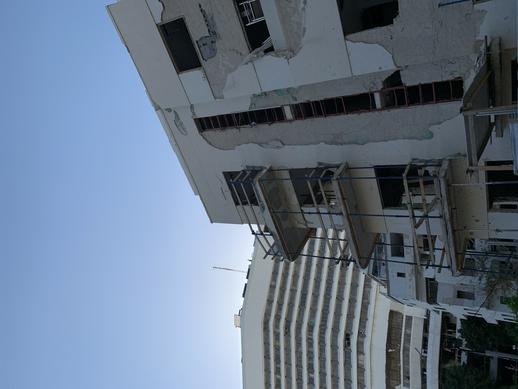 The image depicts a damaged building in Tel Aviv, Israel. The structure shows signs of significant damage, with sections of the exterior facade missing or crumbling. There are open window frames and exposed interior elements, suggesting structural compromise. Scaffolding is visible, indicating ongoing construction or repair efforts. In the background, another tall building is visible, contrasting the damaged building with a more intact structure. The sky is clear and blue. The overall impression is of a site undergoing restoration following damage, possibly from conflict or natural disaster.
