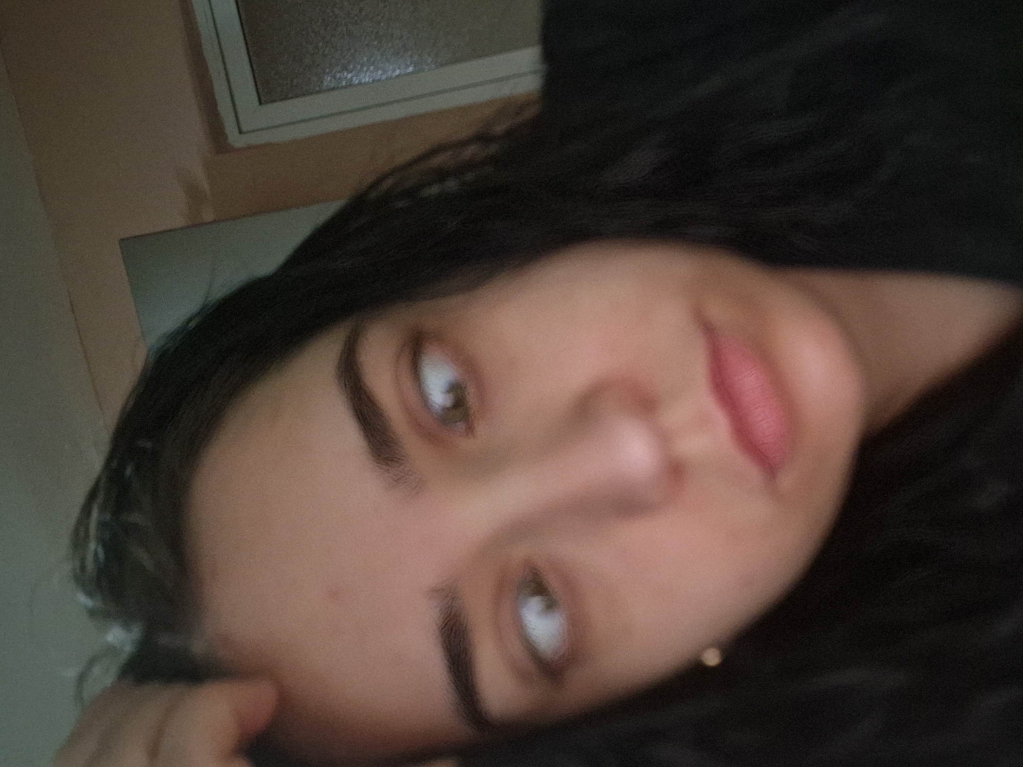 A blurry, close-up, slightly rotated image shows a person with dark hair, light skin, and brown eyes. The person is looking towards the camera. A hand is visible in the lower left corner of the image, suggesting the person may be lying down. There is a window and wall in the background. The image appears to be taken indoors. Unknown, Unknown is not confirmed as a location in this image.
