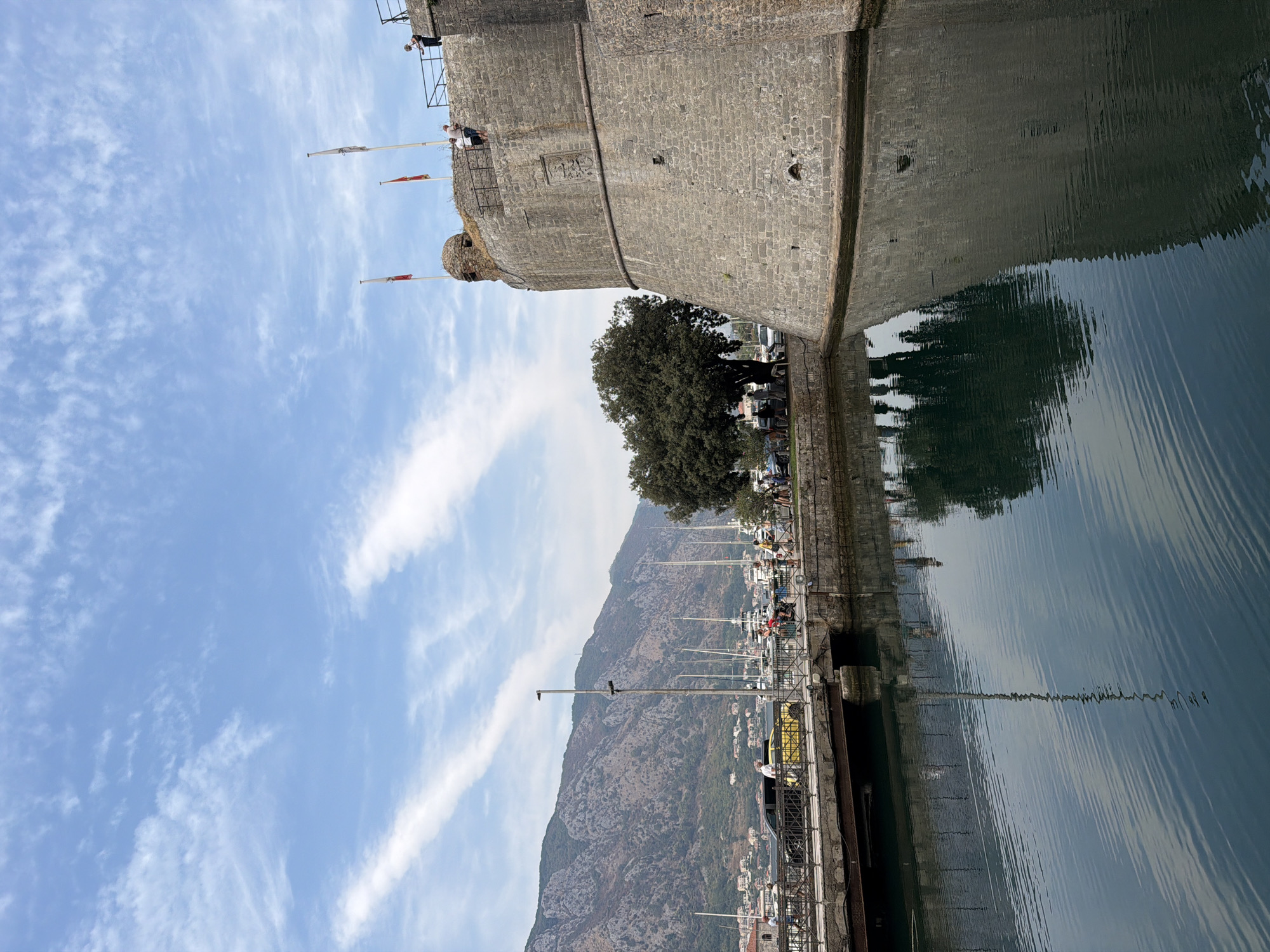 A wide, bright, slightly cloudy sky is visible above a scene that includes a fortified stone structure, water, a bridge, a street with pedestrians and vehicles, sailboats, and a mountain range. The fortified structure, appearing to be a wall or tower of a castle, is constructed of large, rough-hewn gray stones and has a rounded, imposing appearance. Two flags, red with white symbols, fly from poles atop the structure. A few individuals are visible on the parapet of the fortification.
In the foreground, calm, dark blue water fills the bottom portion of the image, reflecting the sky and the fortified structure. A bridge, also appearing to be made of stone and metal, spans the water in the middle ground, connecting two sides of the waterway. A street runs along the edge of the water on the far side of the bridge, with several people walking and what appear to be a few parked cars, including a bright yellow one. Beyond the street, numerous sailboats with tall masts are docked, indicating a harbor.
In the background, a large, verdant mountain range dominates the horizon, its slopes covered in trees and dotted with buildings. The overall impression is of a historical coastal town with a fortified presence overlooking a harbor and natural landscape. The location is the city of Kotor, Montenegro, known for its well-preserved medieval old town and fortifications.