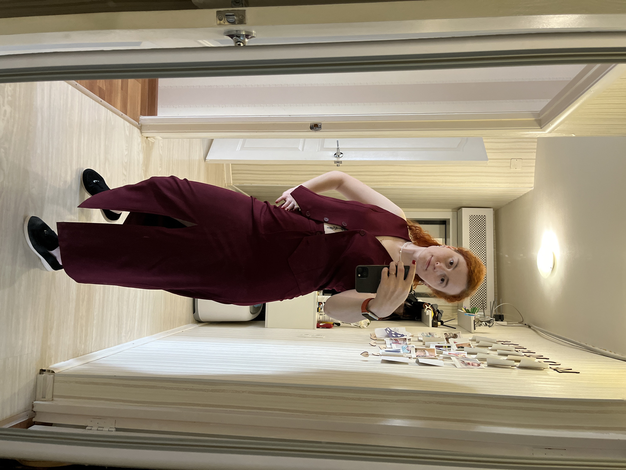 A red-haired woman is taking a selfie in a mirror. She is wearing a burgundy outfit. The location appears to be inside a room with light-colored walls and flooring. There are various items on a surface below the mirror, including cards and other small objects. A radiator can be seen in the background. No information suggests this image was taken in the city of Chaikovsky, Russia.