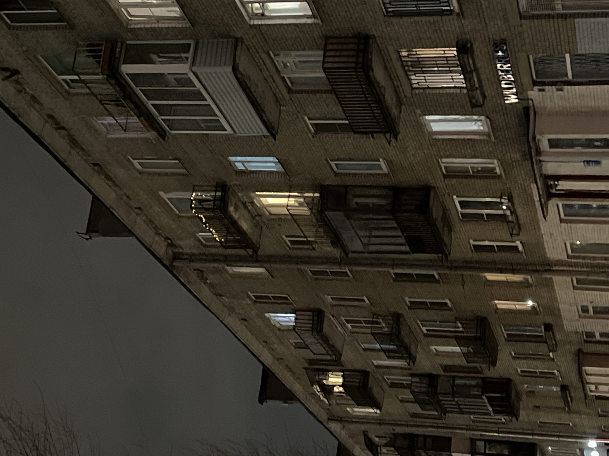 Here's a description of the image:
The image depicts the exterior of a multi-story brick building at night, likely an apartment complex. The building is viewed from a low angle, tilted upwards, with the top of the building appearing to recede into the darkened sky. The building's facade is characterized by rows of windows, some with metal balconies or grates. Some windows are lit, suggesting occupancy.
Notably, the sign "WILDBERRIES" is visible, indicating a commercial presence.
The scene suggests a densely populated urban environment, potentially located in Saint Petersburg, Russia.