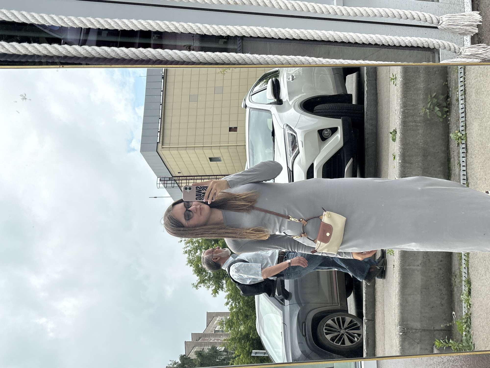 A young woman is taking a selfie in a large mirror, reflecting an outdoor urban environment. She is wearing a long, light grey dress and sunglasses, and carrying a small handbag over her shoulder. In the reflection, behind her, is an older man with a backpack walking past. Two cars are visible: a dark grey SUV in the foreground and a white SUV behind it. The background shows buildings and trees, suggesting a city street. The mirror appears to be a shop window or part of an establishment. This scene is located in the city of Perm, Russia.