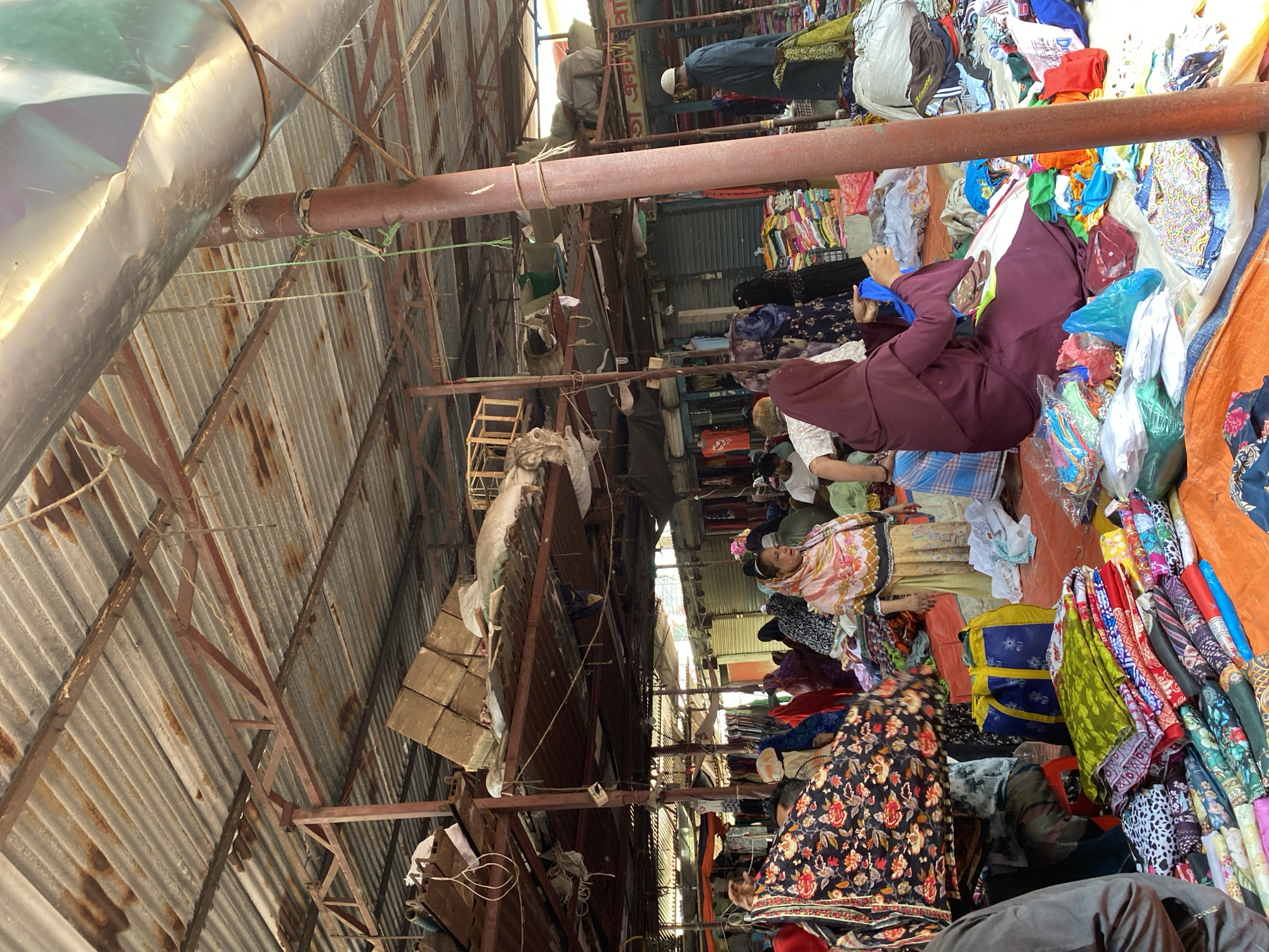 This photograph shows an indoor marketplace in Dhaka, Bangladesh, identified by the corrugated metal roofing and support beams. Several vendors and customers are present. The vendors, some wearing headscarves or traditional clothing, are sitting on the ground, surrounded by piles of fabrics and clothing laid out for sale. Customers are milling about, some looking at the goods. A variety of garments are visible, including colorful saris and various other clothing items. Bags, possibly containing purchased items, are also present. The market area is somewhat crowded, with a variety of items stored overhead. The lighting is dim and uneven.