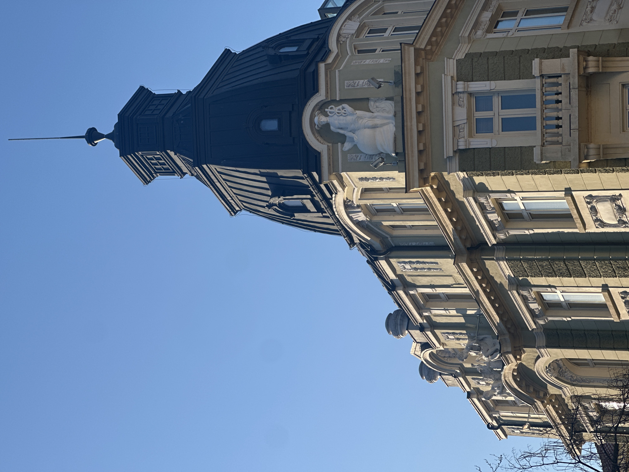 The image shows the architectural detail of a building located in Sofia, Bulgaria. The building features a tall, dark-colored spire atop a multi-tiered roof. The facade of the building is decorated with classical architectural elements, including moldings, columns, and a relief sculpture of a figure. The building is lit by bright sunlight against a clear blue sky.