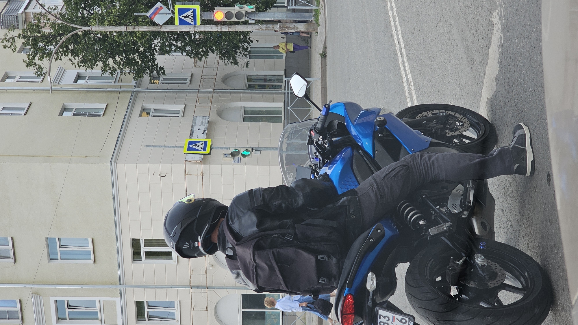 A person wearing a black helmet and a black leather jacket with a black backpack is sitting on a blue motorcycle at an intersection in the city of Kamensk-Uralsky, Russia. The motorcycle is facing away from the camera, with its rear tire visible in the foreground. The person is wearing dark pants and dark sneakers. In the background, there is a light-colored building with several windows. Across the street, there are traffic lights and pedestrian crossing signs. A woman in a yellow shirt and purple pants is standing on the sidewalk. The scene appears to be a city street on a cloudy day.