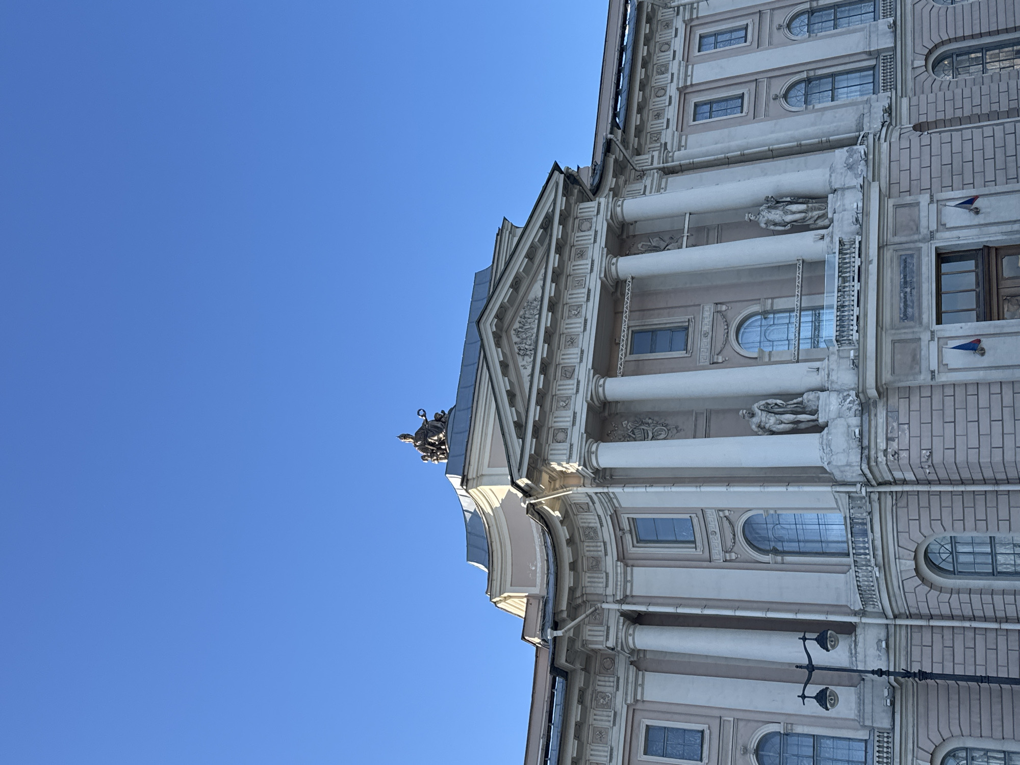 This image depicts a partial view of the Smolny Cathedral in Saint Petersburg, Russia, captured from a low angle. The sky is a clear, bright blue, contrasting with the building's pale facade. The architecture exhibits classical elements such as columns, arched windows, and decorative statues. There is also a sculptural group atop the roofline. A street lamp is visible on the right side of the building. The lighting suggests a sunny day.
