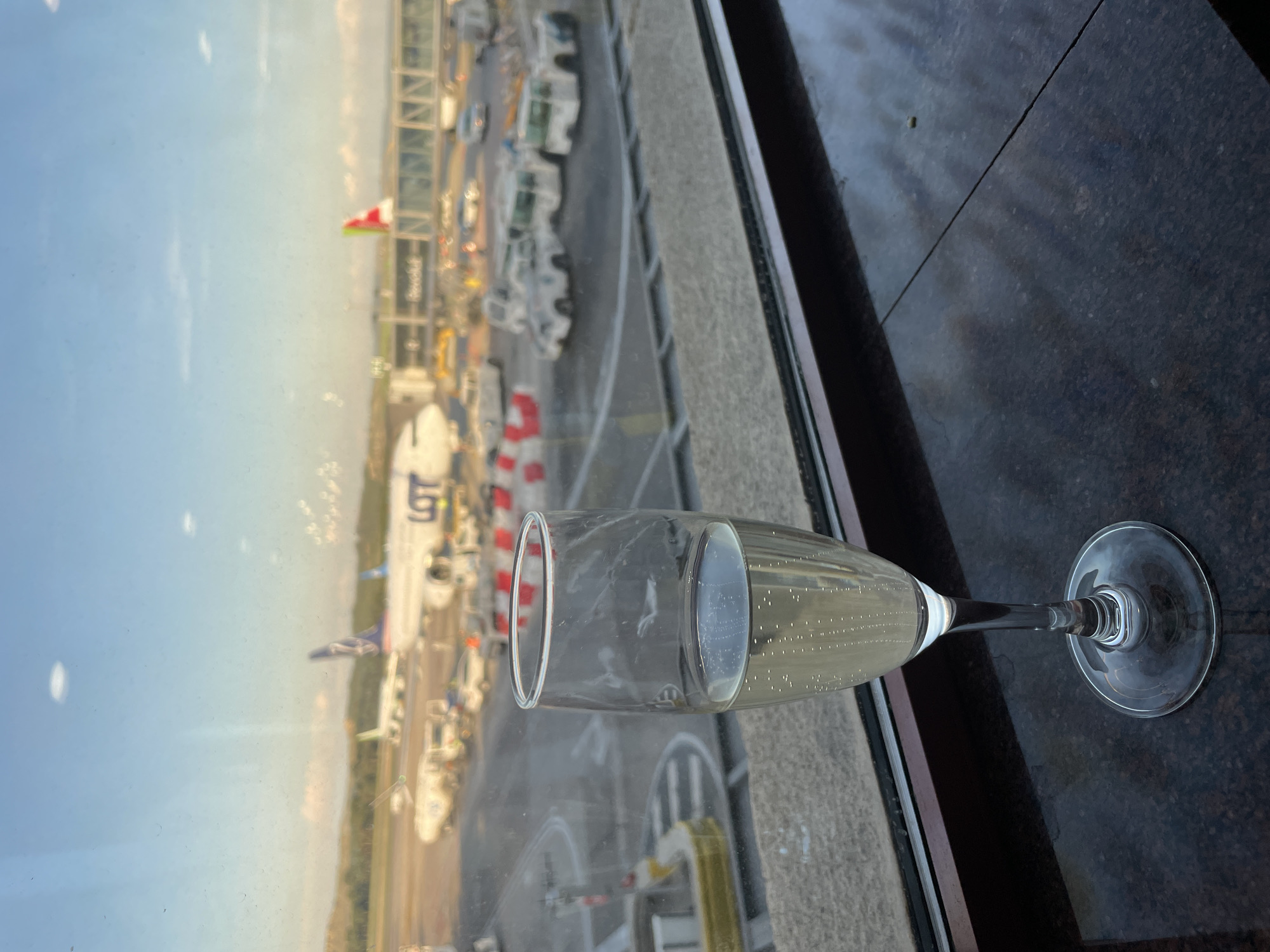 An aerial view of Madrid Barajas Airport is visible through a window with a champagne flute in the foreground. The champagne flute is partially filled with a bubbly liquid. Outside, the airport is bustling with activity, including parked planes and various airport vehicles. The sky is clear.