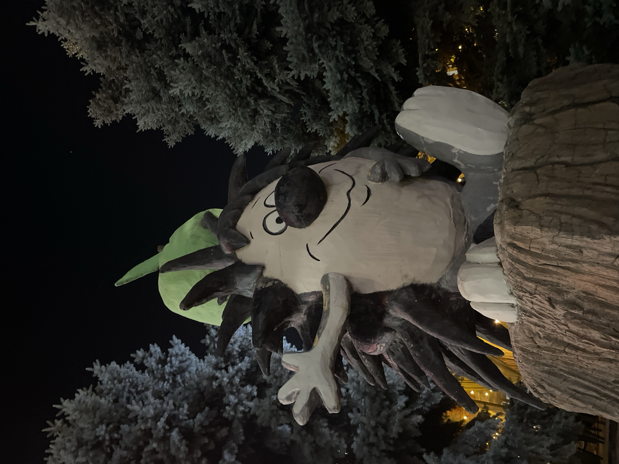 An outdoor, eye-level, medium shot captures a whimsical, wooden sculpture of a cartoonish hedgehog character at night in the city of Almaty, Kazakhstan. The hedgehog, rendered in muted colors of gray, white, black, and green, sits atop a textured, brown, wood-like base, appearing to be made of carved and painted wood. Its large, round, dark nose and wide, smiling mouth convey a friendly expression, while its eyes are depicted as black circles with white pupils. The character has several dark, pointed quills protruding from its back, and a large, green, leaf-like protrusion on its head. One of its arms is raised and extended forward, with a five-fingered hand. Its legs are bent, with large, rounded feet resting on the base. The hedgehog is surrounded by tall, dark green evergreen trees, whose branches are visible against the black night sky. Subtle, warm yellow lights are visible in the background, suggesting artificial lighting or decorations within the park or public space. The lighting on the sculpture highlights its form and details.