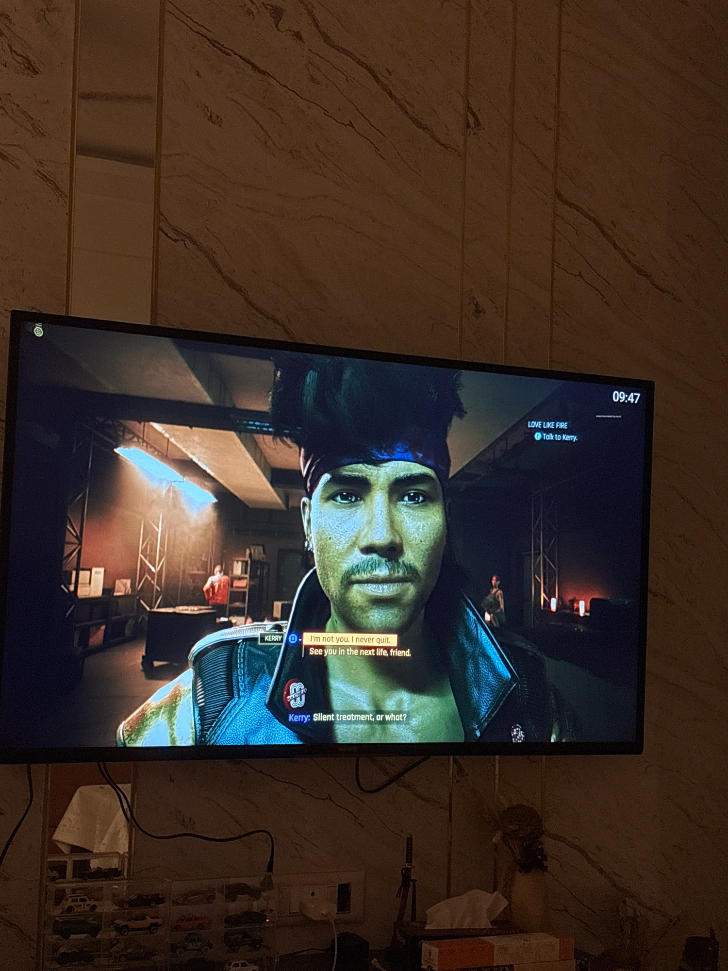 A television screen displays a video game scene featuring a close-up of a male character's face with a bandana and a leather jacket. The character's name, "Kerry," is visible in a subtitle box. Another subtitle reads, "I'm not you. I never quit. See you in the next life, friend." In the background of the game, another person in an orange jacket is visible, along with bookshelves and stage lighting. The game's interface shows the time as "09:47" and a mission name "LOVE LIKE FIRE." The television is mounted on a wall with a marble-like textured finish. Below the television, a collection of miniature toy cars is displayed on shelves. A tissue box and other items are on a table in the foreground. The scene is illuminated by ambient light from the television and possibly room lighting, creating shadows and highlights on the wall and objects. There is no visible indication of the specific location within Bengaluru, India.