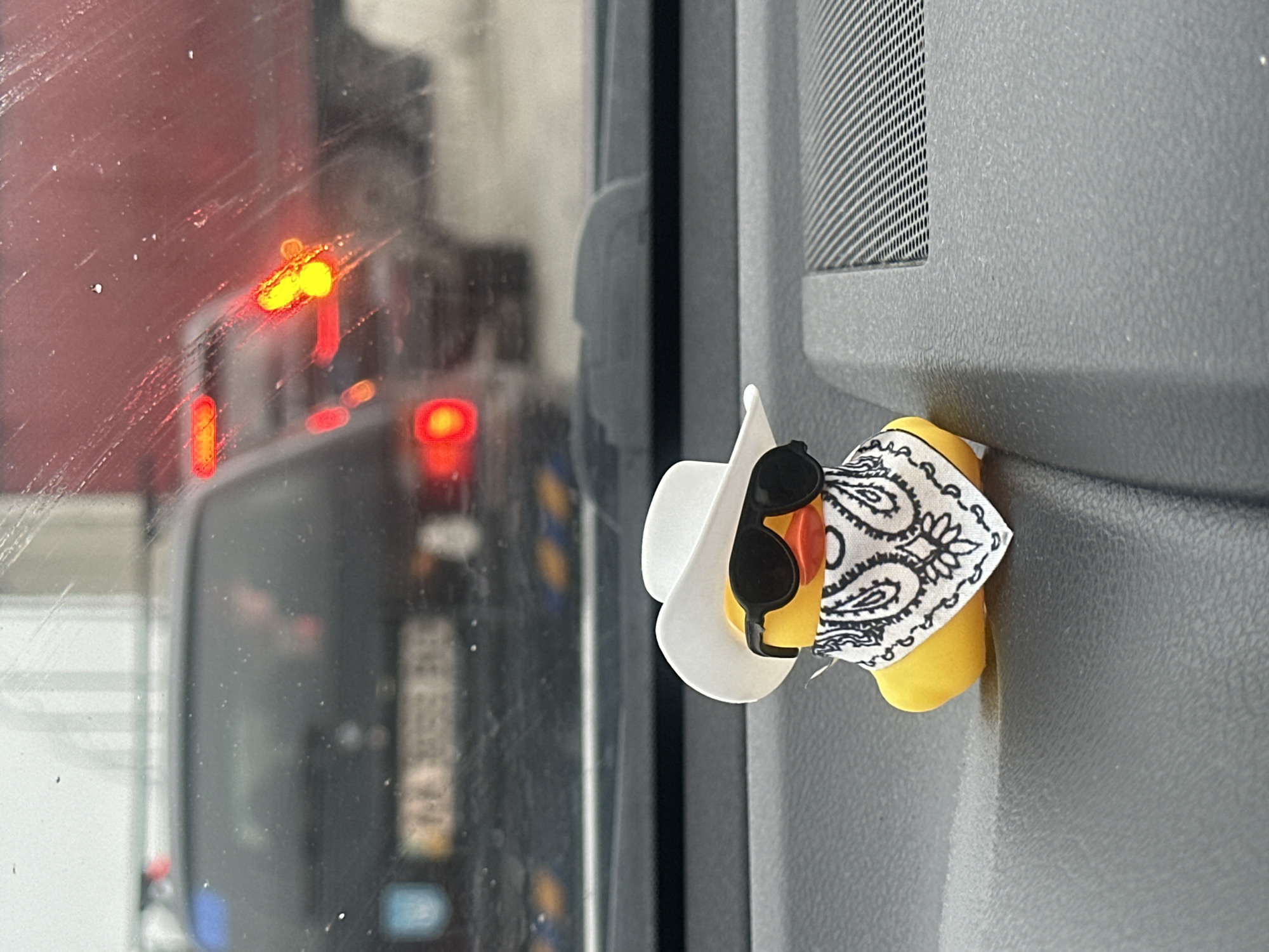 The image depicts a yellow rubber duck attached to the interior of a vehicle, possibly a van or truck. The duck is wearing a white cowboy hat, black sunglasses, and a white bandana with a black paisley pattern. Through the vehicle's window, traffic is visible, including the rear of a truck or bus with blurred tail lights. The surroundings are suggestive of an urban environment. There is no evidence in the image to indicate it was taken in Kyiv, Ukraine.