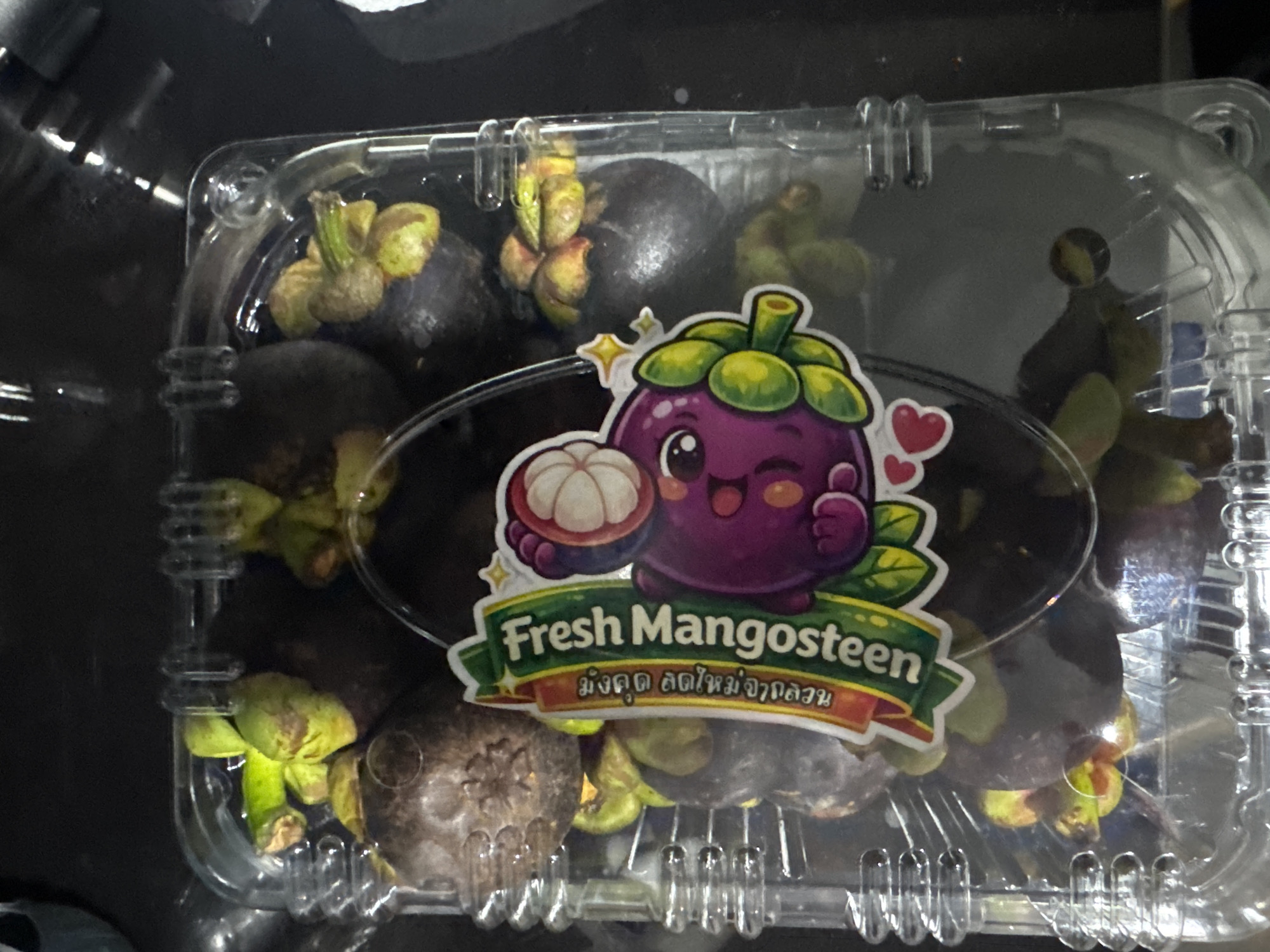 A clear plastic rectangular container, filled with approximately a dozen dark purple mangosteens (Garcinia mangostana), is centrally positioned on a dark, indistinct surface. The container has ribbed sides for structural integrity and a slightly domed lid. A colorful, die-cut sticker label is affixed to the center of the lid by a thin, clear plastic strap. The label features a green banner with the English text "Fresh Mangosteen" in white font. Below this, Thai script reads "มังคุดสดคุณภาพดี," translating to "Fresh Quality Mangosteen." To the right of the text is a stylized, smiling purple cartoon mangosteen character with large white eyes, a green calyx, and a cross-section revealing its white internal aril. Two small pink hearts are depicted near the character's base. The mangosteens inside are mostly dark purple with green-to-brown star-shaped calyxes visible at their tops. The background is uniformly dark and out-of-focus, suggesting an indoor setting such as a kitchen counter or a market stall. The presence of Thai script on the product label strongly indicates that this scene is located in Thailand, with Bangkok being a plausible city for such commercially packaged fruit to be sold or photographed.